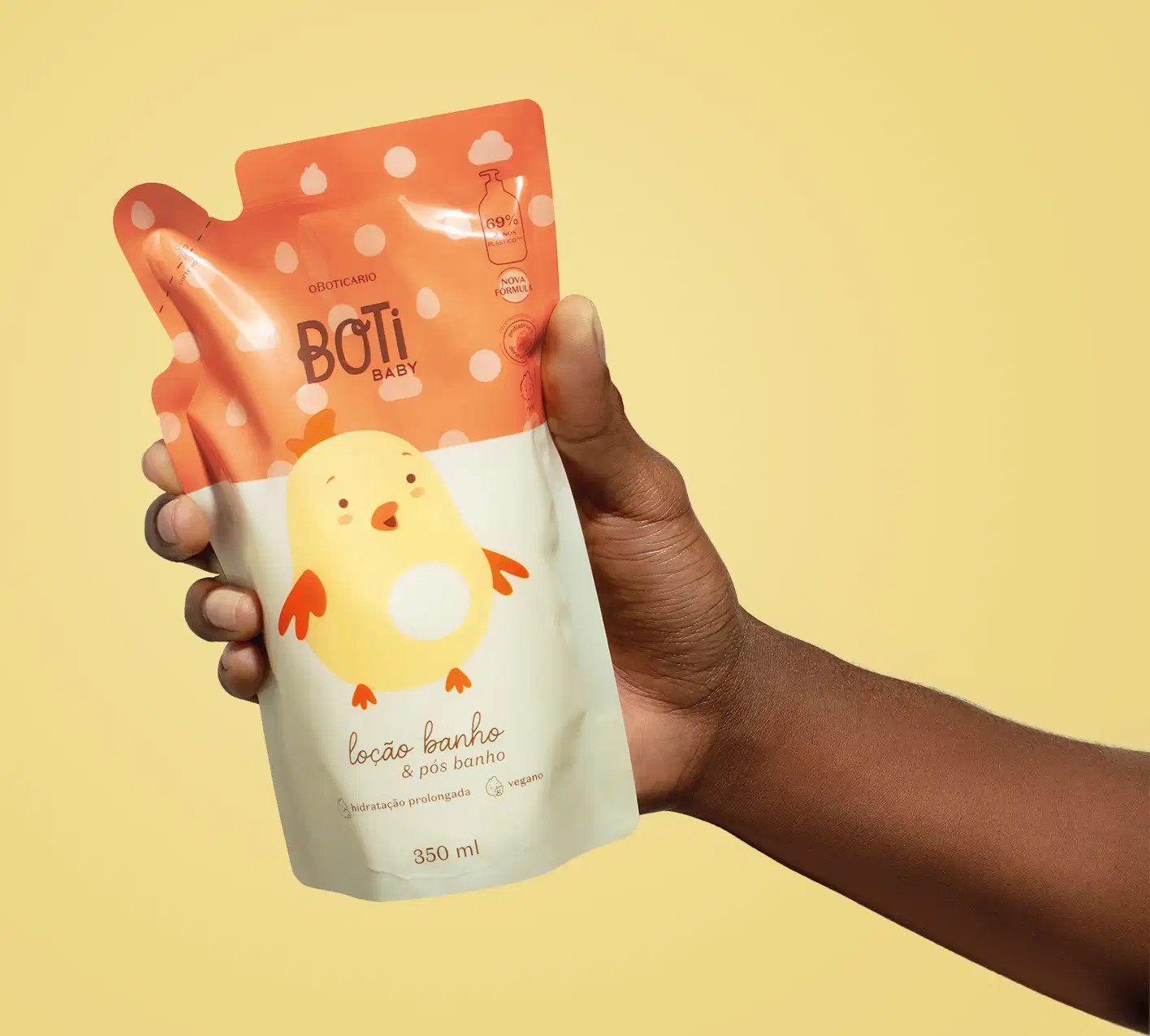 Mão femininas segurando um refil de loção hidratante corporal da marca Boti Baby em forma de sachê nas cores laranja e amarelo claro com ilustração de passarinho amarelo com detalhes laranjas. O fundo da imagem é laranja claro.
