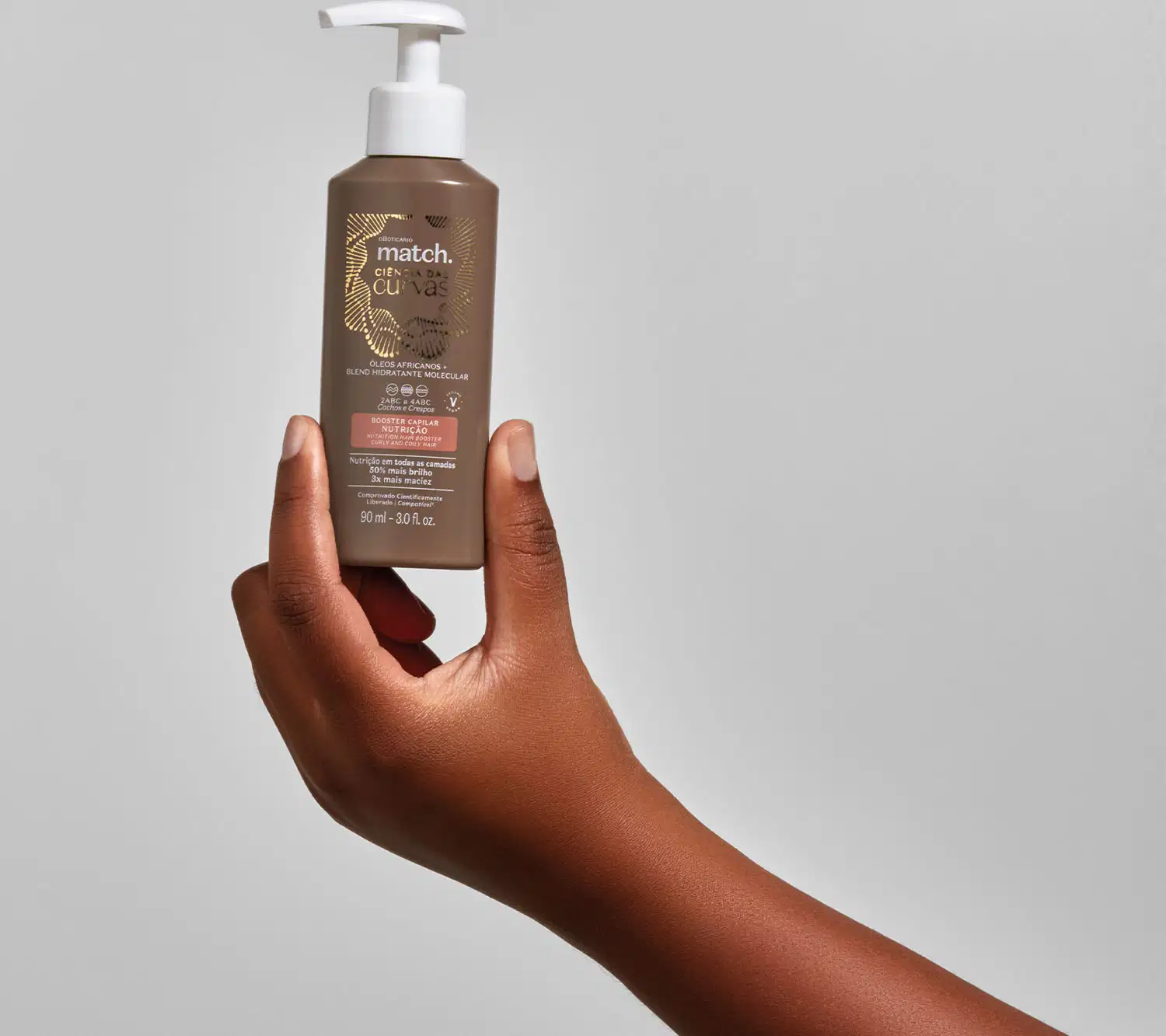 Mão segurando um frasco marrom com bomba dosadora branca e letras brancas e bege claro contendo 90ml de booster capilar Nutrição Match Ciência das Curvas em fundo cinza.