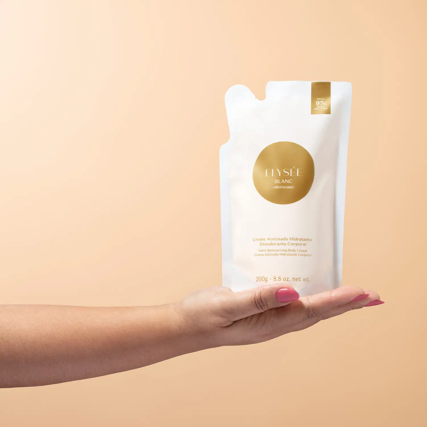 Mão feminina segurando um refil de creme desodorante hidratante corporal acetinado da marca Elysée Blanc em sachê de 200g nas cores branca e dourada, em fundo em tons de rosa.