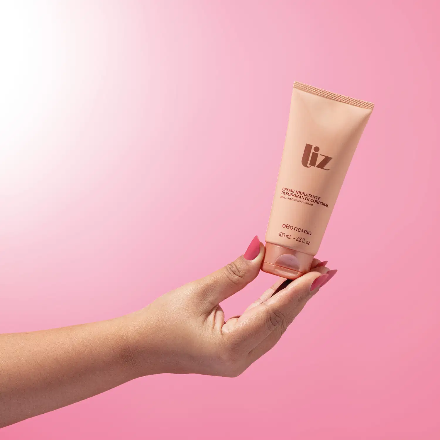 Mão segurando uma bisnaga rosa com letras marrons de creme desodorante hidratante corporal da marca Liz em fundo em tons de rosa.