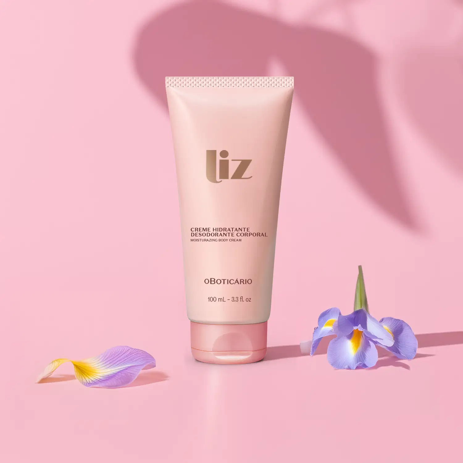 Bisnaga rosa om letras marrons de creme desodorante hidratante corporal da marca Liz em fundo rosa com sombras e flores roxas espalhadas ao redor da embalagem.