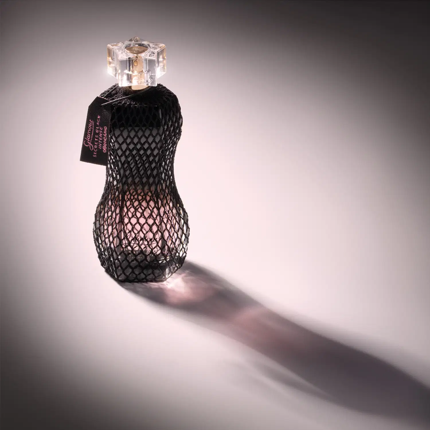 Vidro curvilíneo com renda preta em volta, pulverizador dourado e tampa transparente de desodorante colônia Glamour Secrets Black Intense em fundo claro com a sombra o frasco à frente do item.