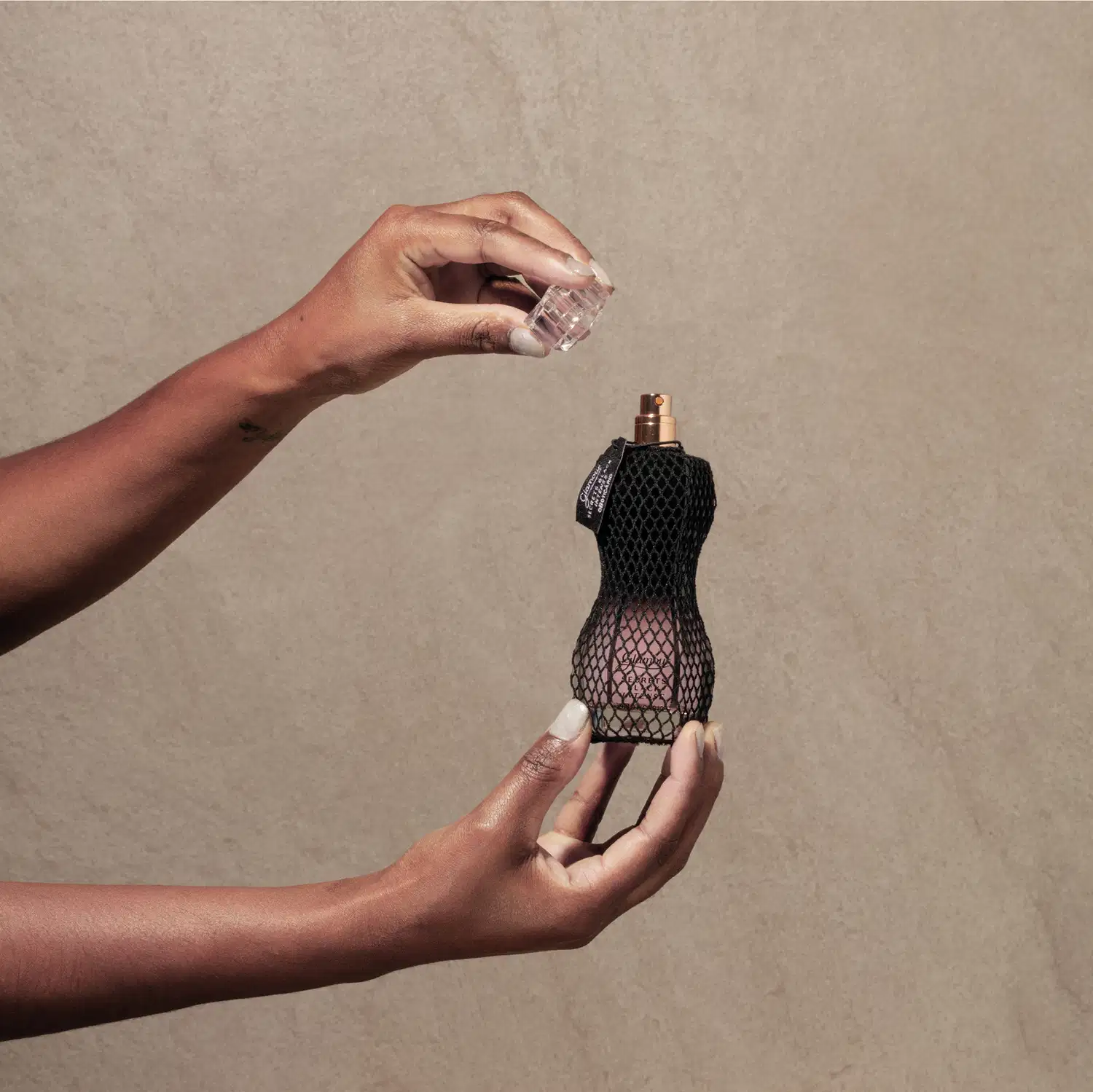 Mão segurando um vidro curvilíneo com renda preta em volta, pulverizador dourado e tampa transparente de desodorante colônia Glamour Secrets Black Intense. A outra mão segura a tampa transparente do produto, tudo em fundo roxo.