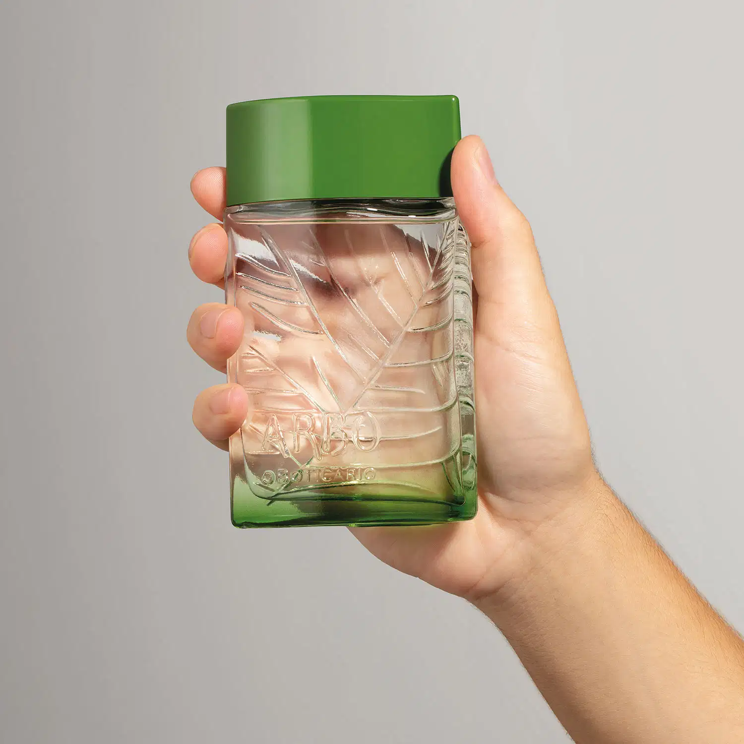 Mão segurando um frasco verde transparente com líquido claro, folhagens em relevo e tampa verde escuro contendo desodorante colônia Arbo Puro em fundo cinza.