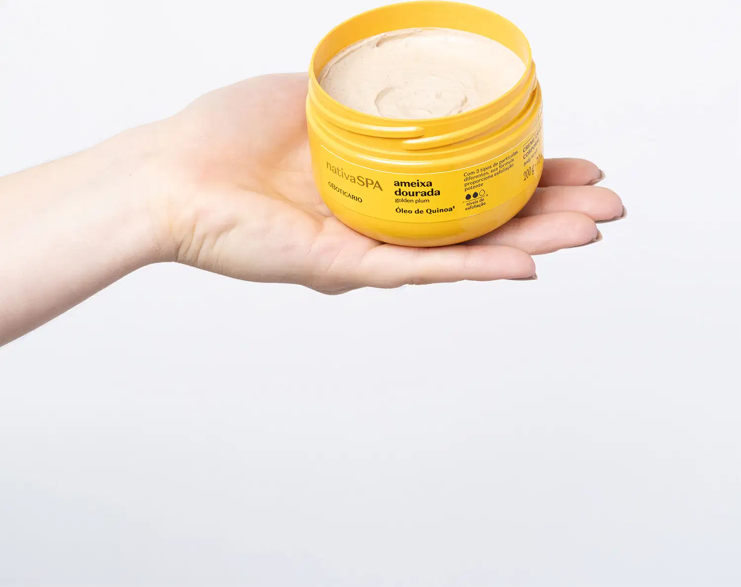 Mão segurando um pote amarelo com tampa e letras pretas contendo creme esfoliante corporal Nativa SPA Ameixa Dourada em fundo azul claro.