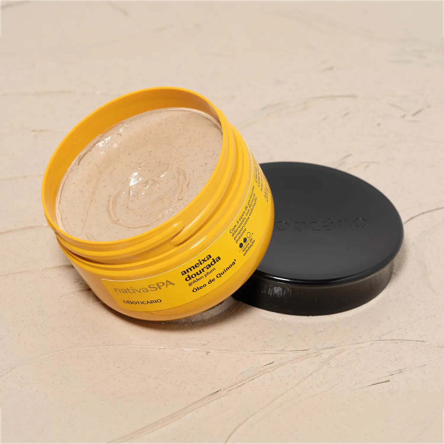 Pote amarelo com letras pretas aberto, mostrando o creme amarelado dentro dele, com a tampa preta ao lado, em superfície bege simulando mármore.