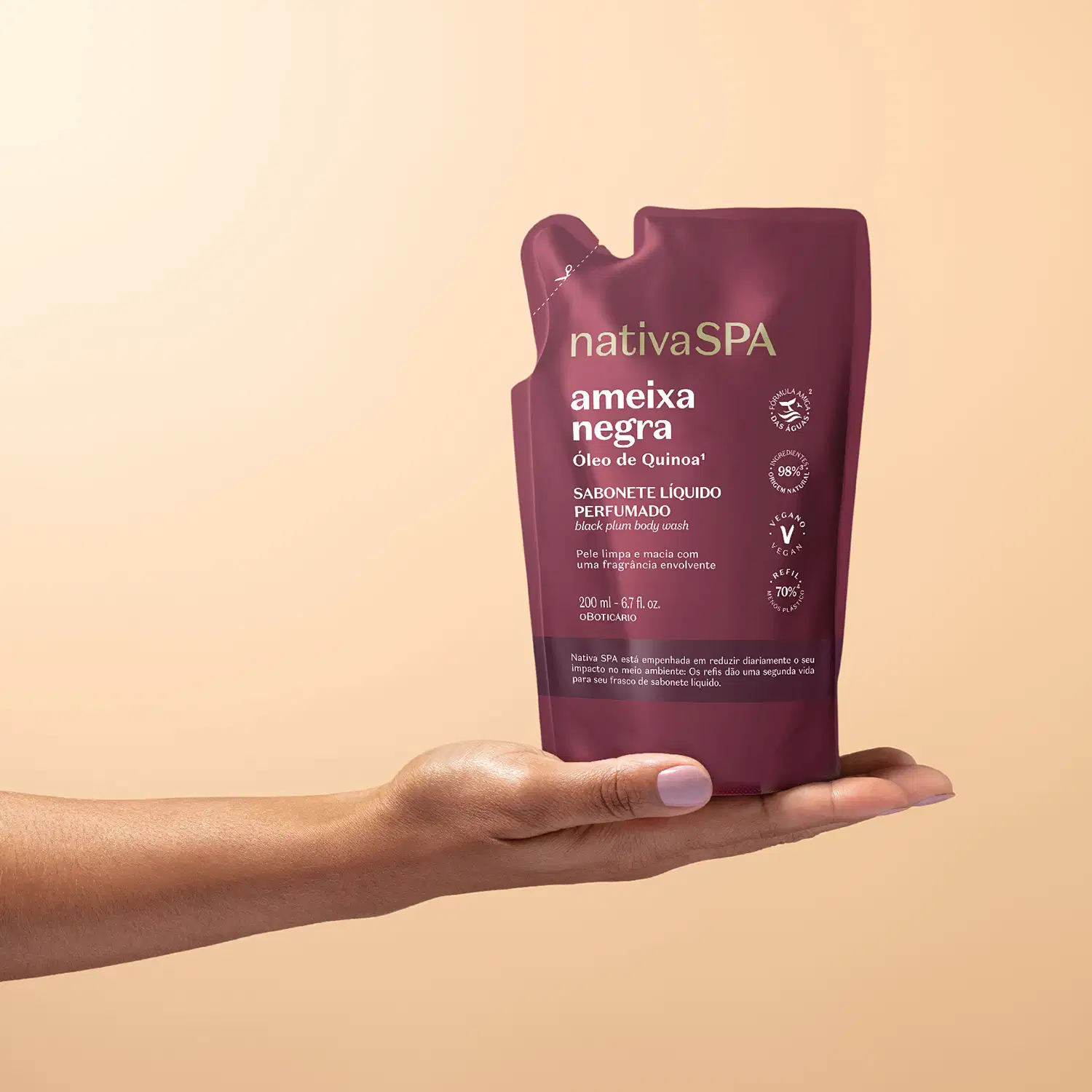 Mão feminina segurando um refil de sabonete líquido perfumado Nativa Spa Ameixa Negra em sachê de 200ml na cor vermelho escuro com detalhe marrom e letras brancas em fundo em tons de rosa.