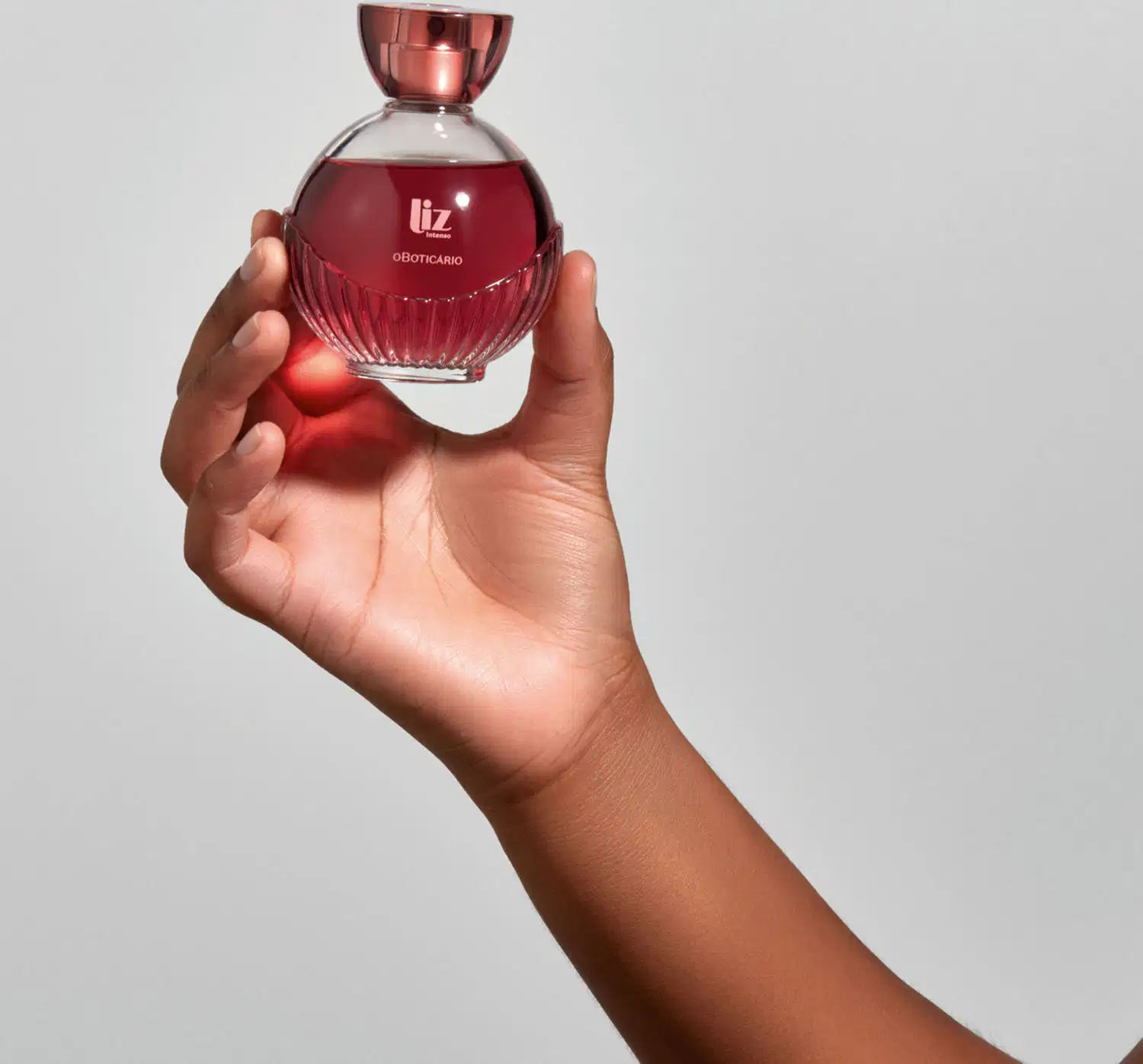 Uma mão está segurando um vidro com líquido vermelho, borrifador dourado, tampa transparente e letras douradas de desodorante colônia Liz Intenso em fundo cinza.