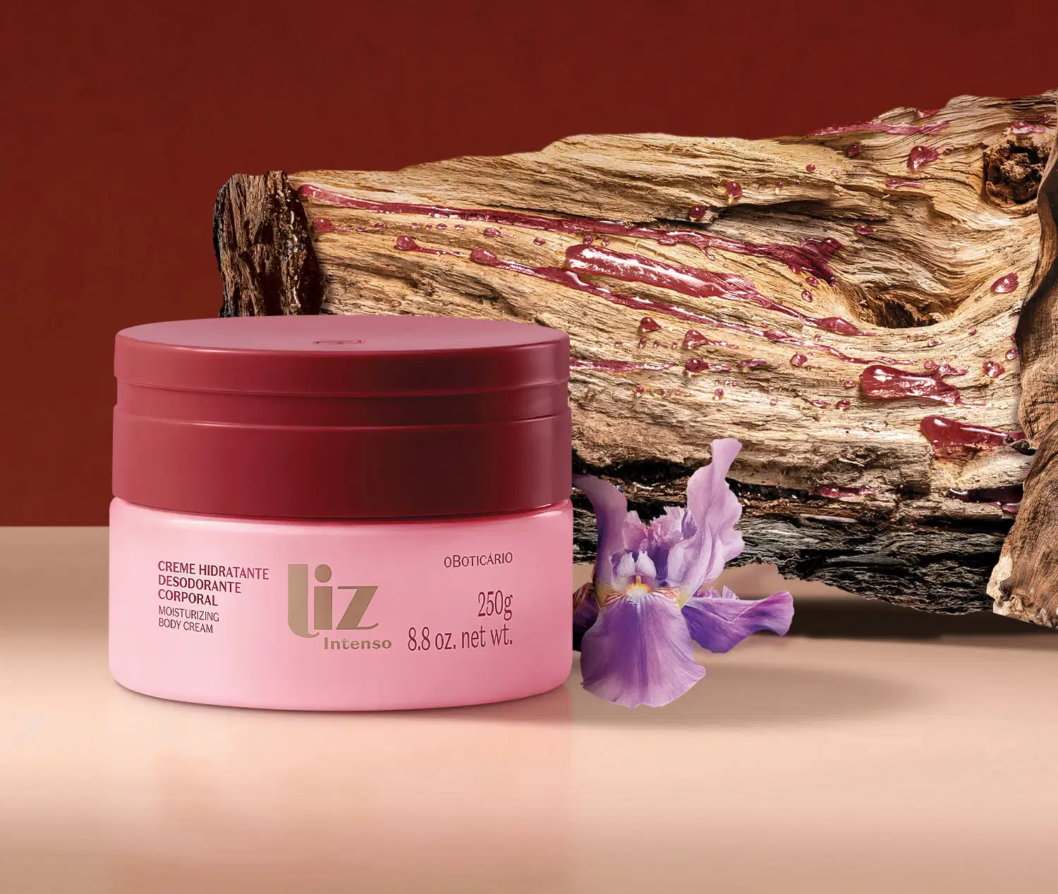 Pote rosa com tampa e letras vermelhas de creme hidratante corporal Liz Intenso ao lado de uma flor roxa e de um galho de árvore, em superfície rosa e fundo vermelho.