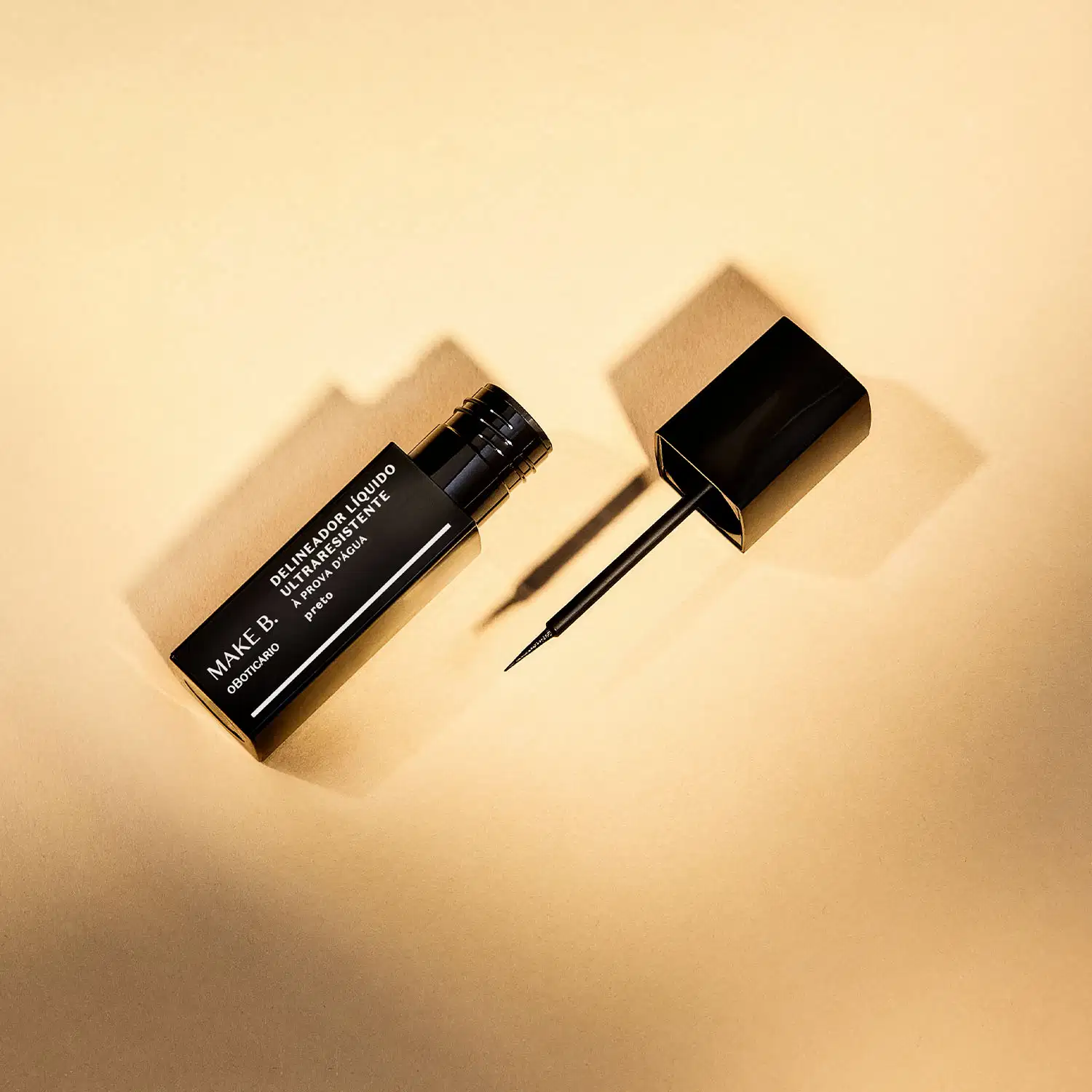 Tubo preto  com letras brancas de delineador líquido para olhos na cor preta da marca Make B., com tampa, haste e aplicador fino. A imagem tem um fundo em tons de rosa.