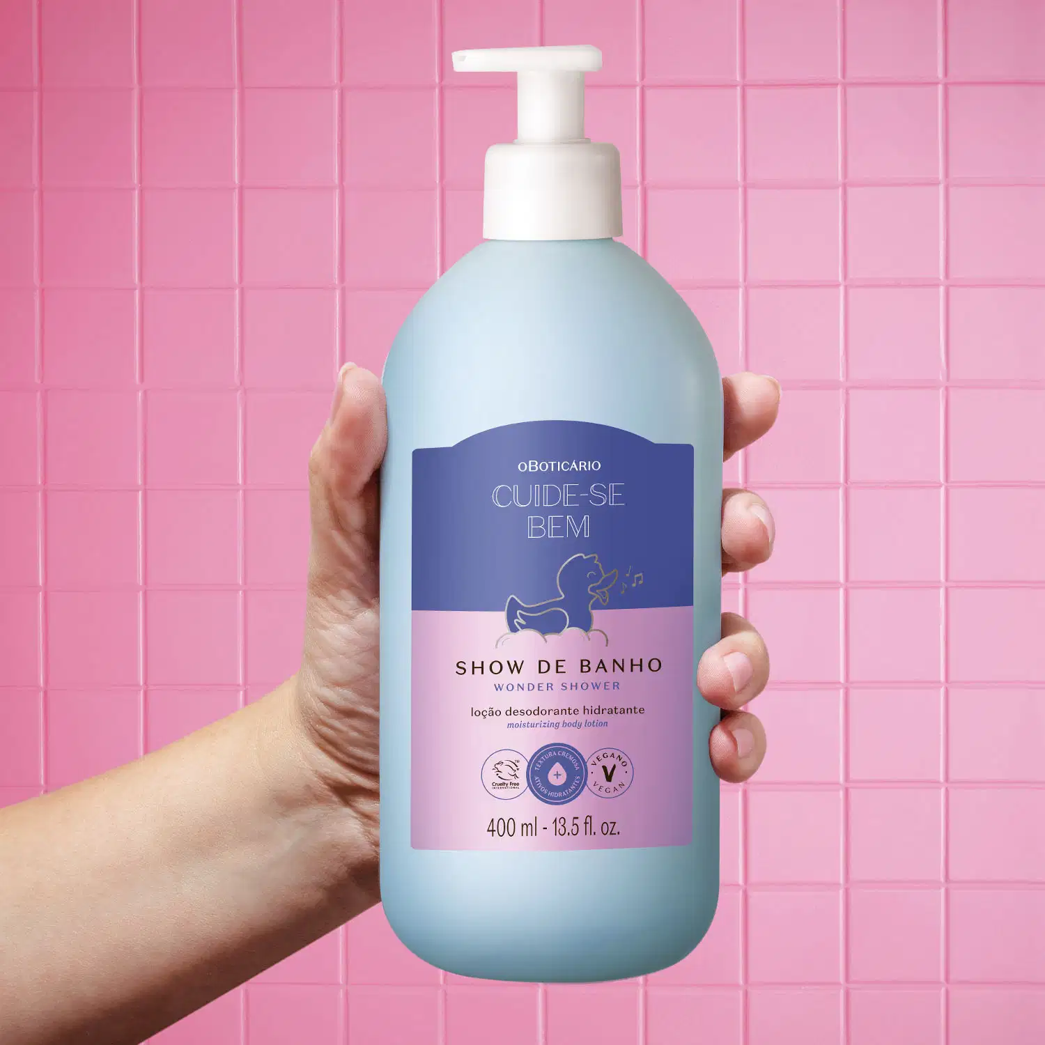 Mão feminina segurando um frasco branco com bomba dosadora e rótulo azul e lilás com ilustração de um patinho de banho de loção hidratante corporal da marca Cuide-se Bem Show de Banho em fundo rosa simulando azulejos de banheiro.