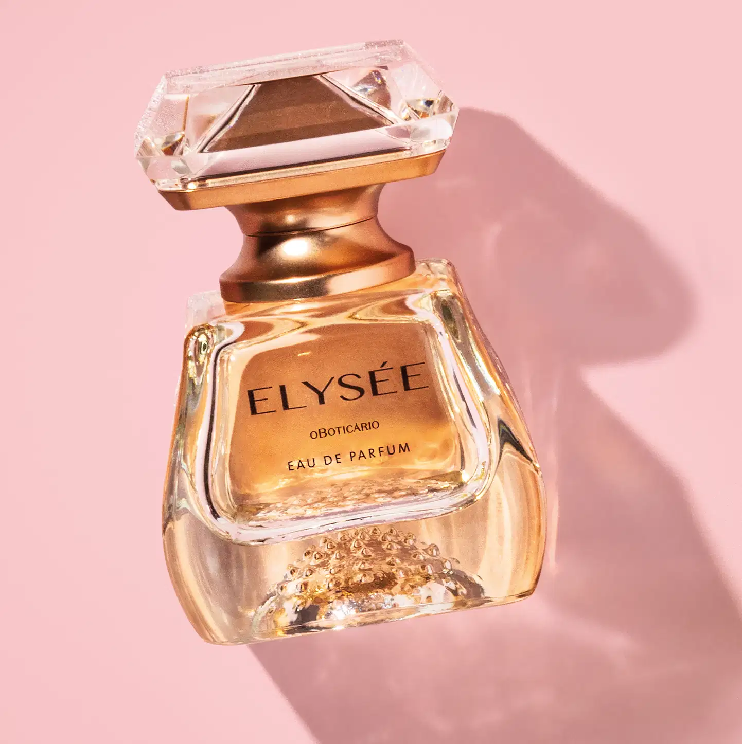 Frasco de Eau De Parfum da marca Elysée em formato geométrico com líquido na cor âmbar e tampa em forma de cristal lapidado, em tons de dourado em fundo rosa claro com sombras.