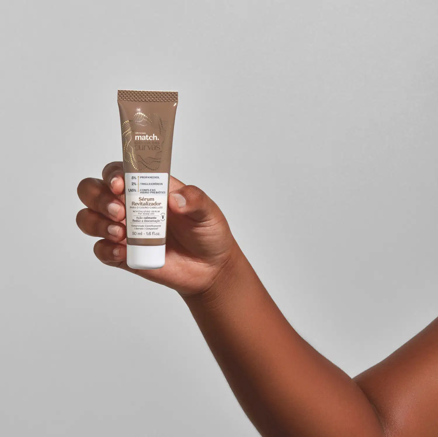 Mão segurando um Frasco marrom com tampa branca, rótulo cinza e letras em bege e preto contendo 50ml de sérum revitalizador para o couro cabeludo da marca Match Ciência das Curvas em fundo cinza.