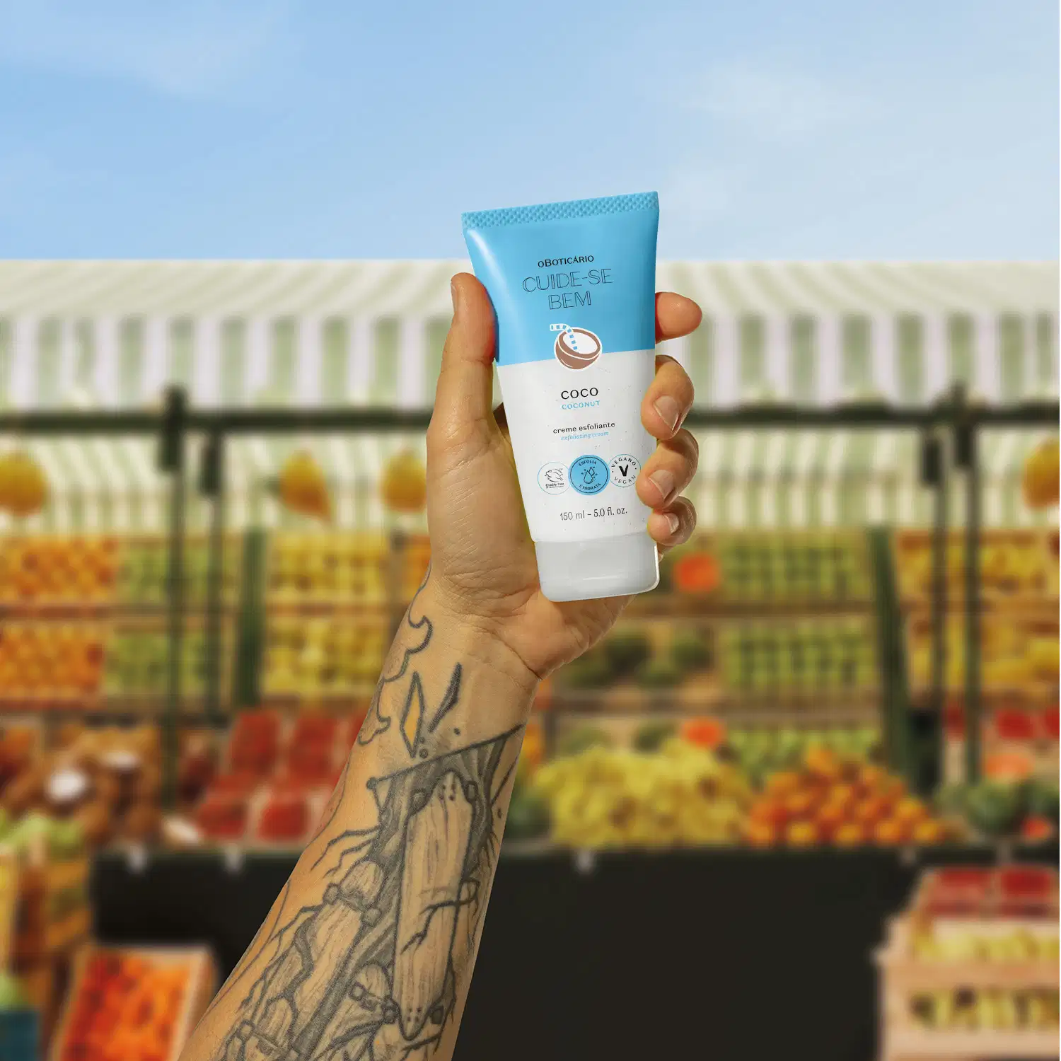 Braço tatuado com mão segurando uma bisnaga azul e branca com coco ilustrado contendo creme esfoliante corporal Cuide-se Bem Feira Coco em um fundo desfocado mostrando uma feira livre.