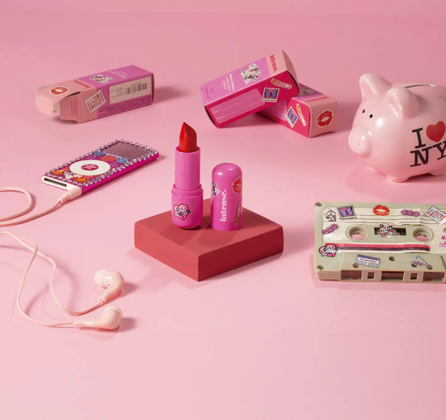 Batom do Arraso Pôr do Sol na cor vermelha da marca Intense em embalagem na cor roxa está em cia de uma plataforma rosa com caixa do produto, mp3 player, cofre e fita cassete, tudo em tons de rosa.