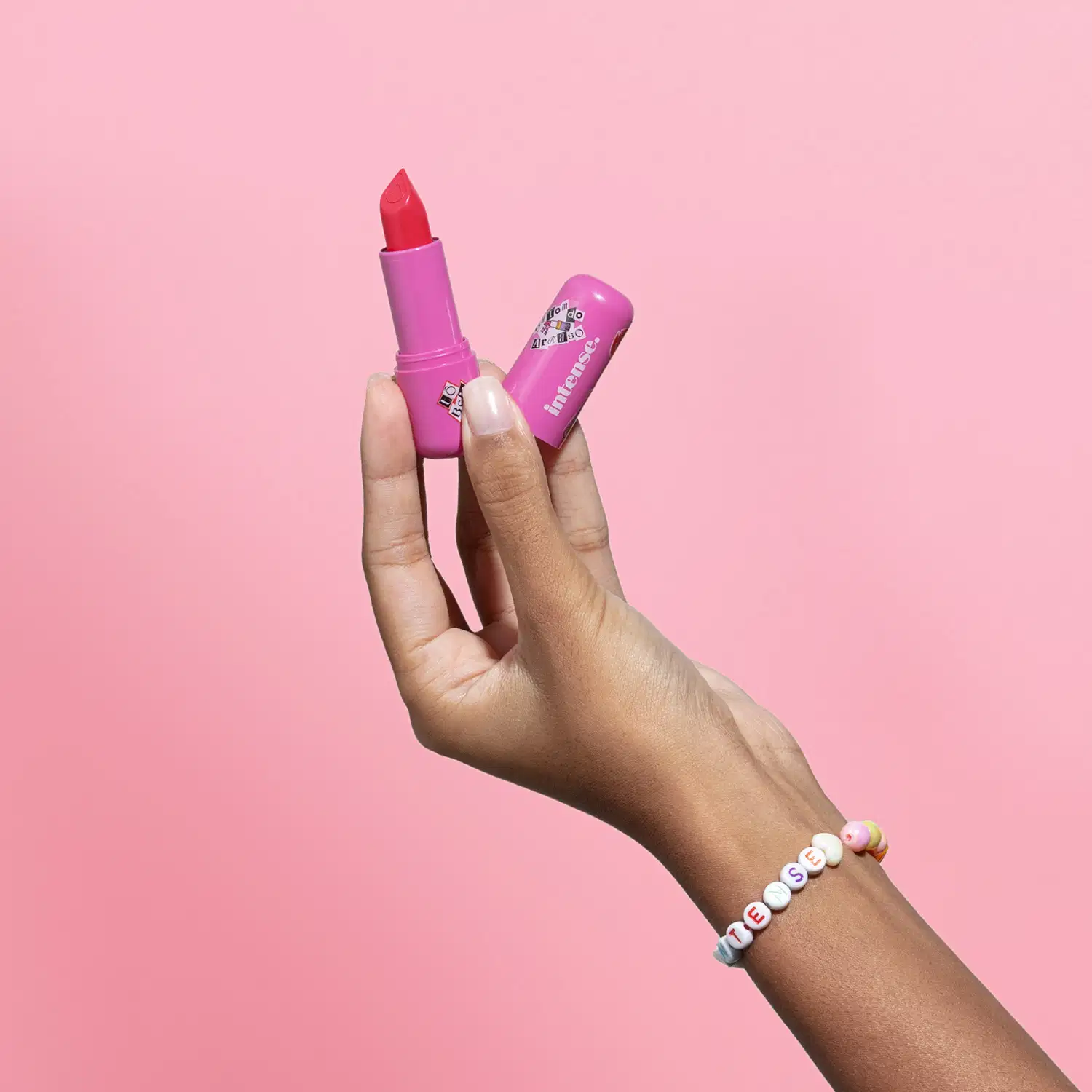 Uma mão feminina segura o batom do Arraso Eu Sou Mais  Eu na cor rosa da marca Intense em embalagem na cor roxa e a outra mão segura a tampa, na mesma tonalidade, em fundo em tons de rosa.