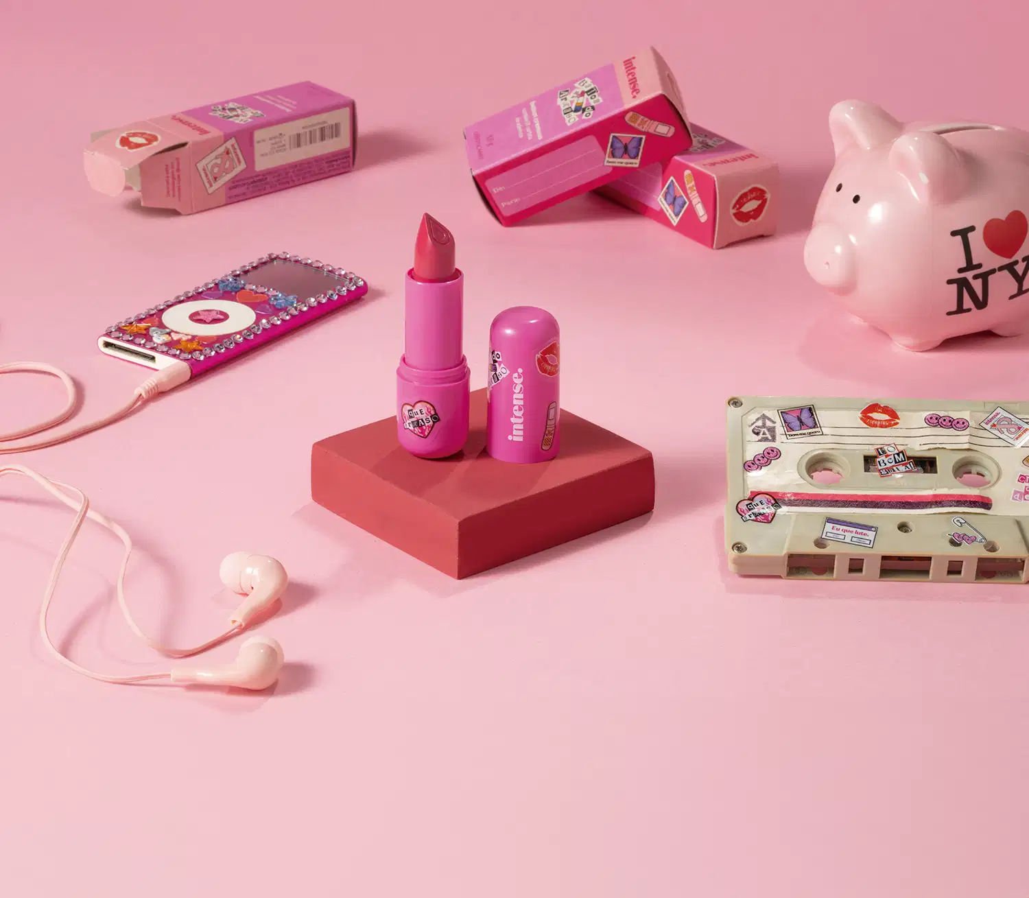 Batom do Arraso Eu Sou Mais  Eu na cor rosa da marca Intense em embalagem na cor roxa está em cia de uma plataforma rosa com caixa do produto, mp3 player, cofre e fita cassete, tudo em tons de rosa.