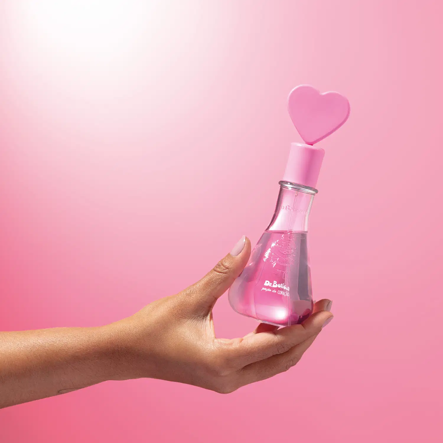 Mão feminina segurando um frasco transparente em formato de vidro laboratorial com liquido rosa, tampa também rosa com coração na ponta e letras brancas no rótulo contendo colônia infantil Dr. Botica Poção do Coração em fundo em tons de rosa.