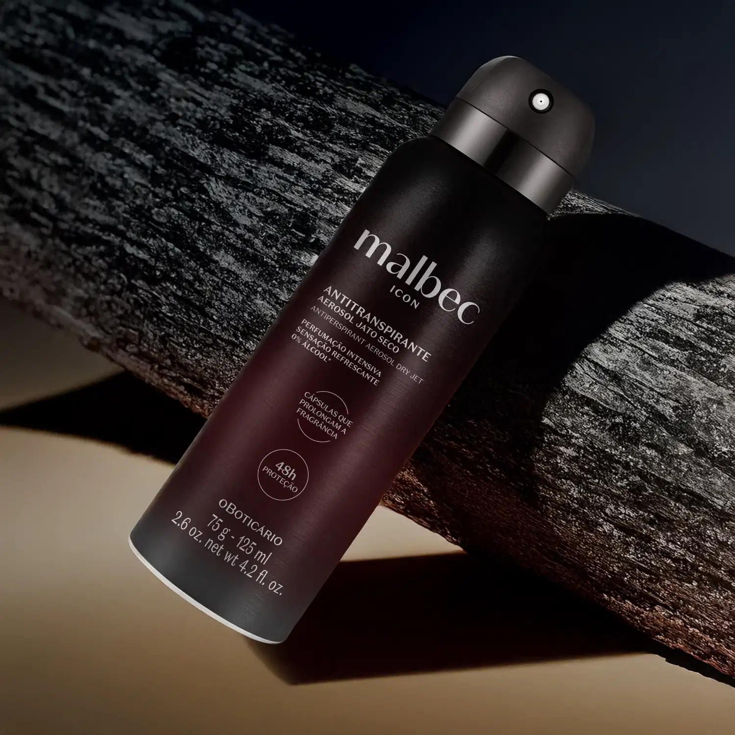 Frasco preto fosco com letras cinzas de antitranspirante aerossol jato seco da marca Malbec Icon sobre superfície de madeira escura, com fundo desfocado e iluminação dramática.