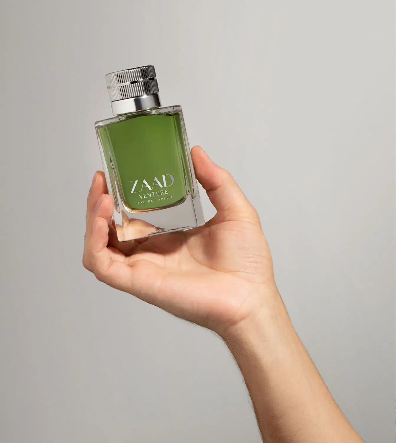 Mão segurando um  vidro retangular na cor verde oliva com tampa texturizada prateada e letras cinza contendo Eau de Parfum Zaad Venture em fundo cinza.