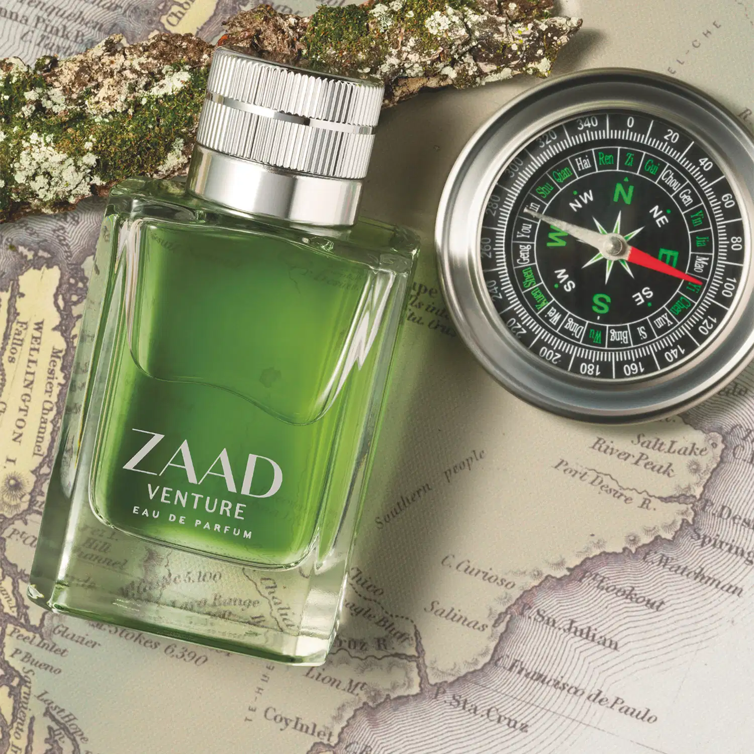 Vidro retangular na cor verde oliva com tampa texturizada prateada e letras cinza contendo Eau de Parfum Zaad Venture em verde, sobre mapa antigo com bússola, em ambiente com musgo e rochas.