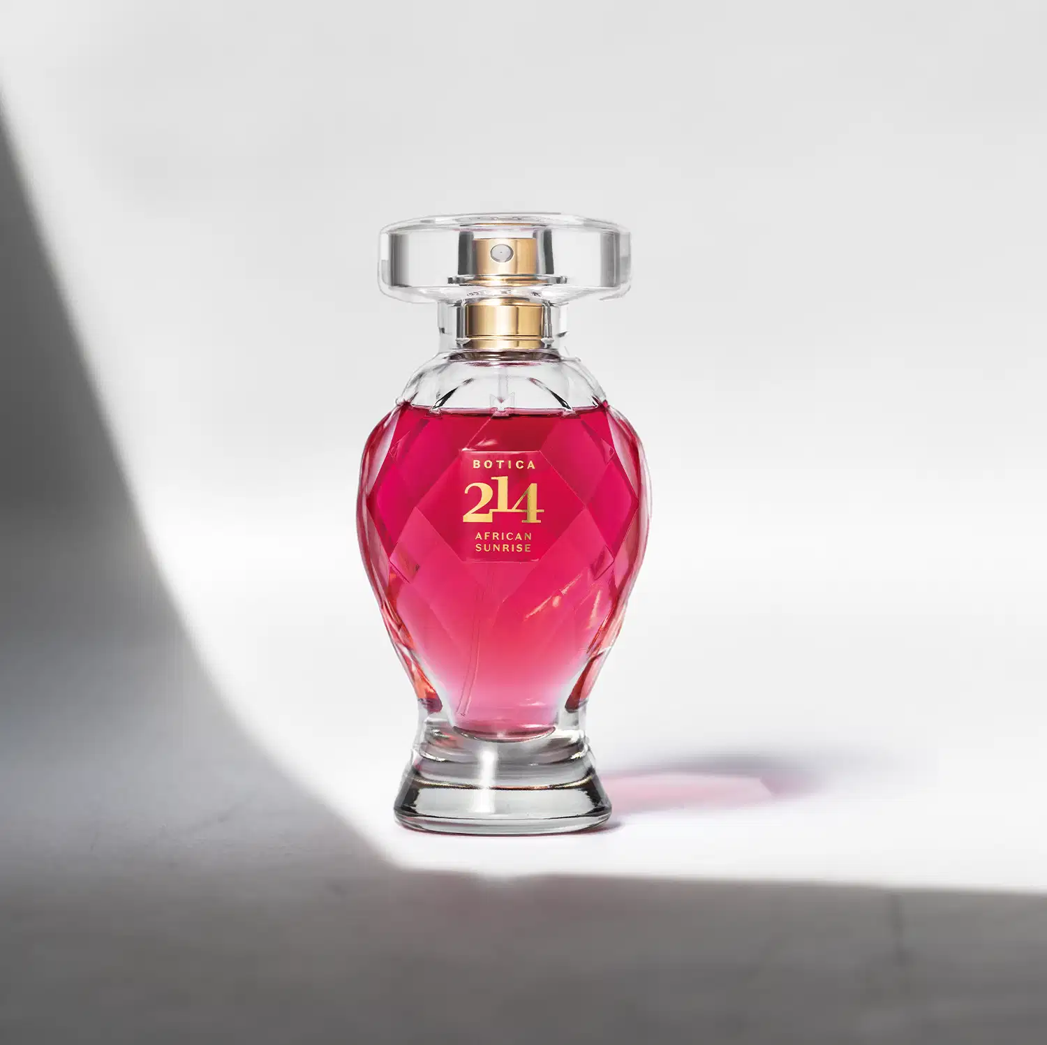 Vidro lapidado em formato de taça com líquido rosa vibrante, tampa transparente, pulverizador e letras douradas contendo 75ml de Eau de Parfum Botica 214 African Sunrise Floral Frutal em fundo claro com sombras.