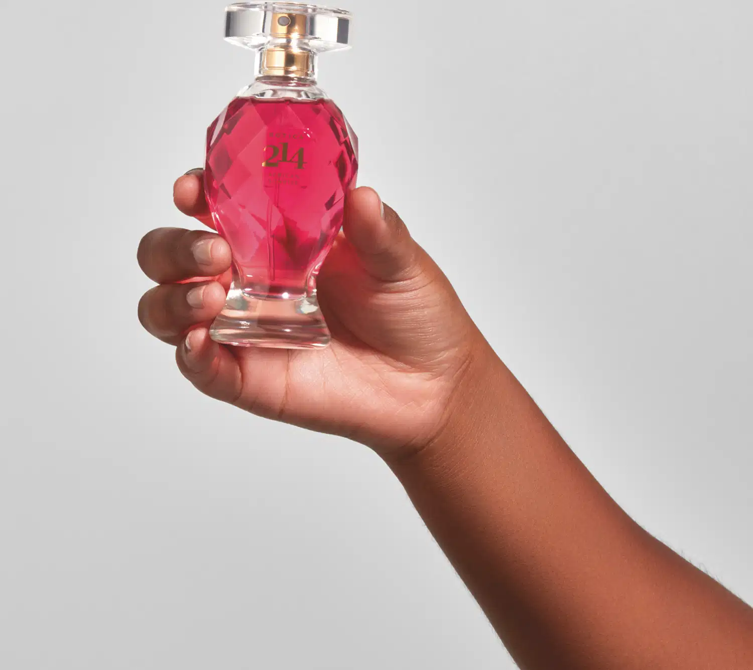 Mão feminina segurando um vidro lapidado em formato de taça com líquido rosa vibrante, tampa transparente, pulverizador e letras douradas contendo 75ml de Eau de Parfum Botica 214 African Sunrise Floral Frutal em fundo azul.