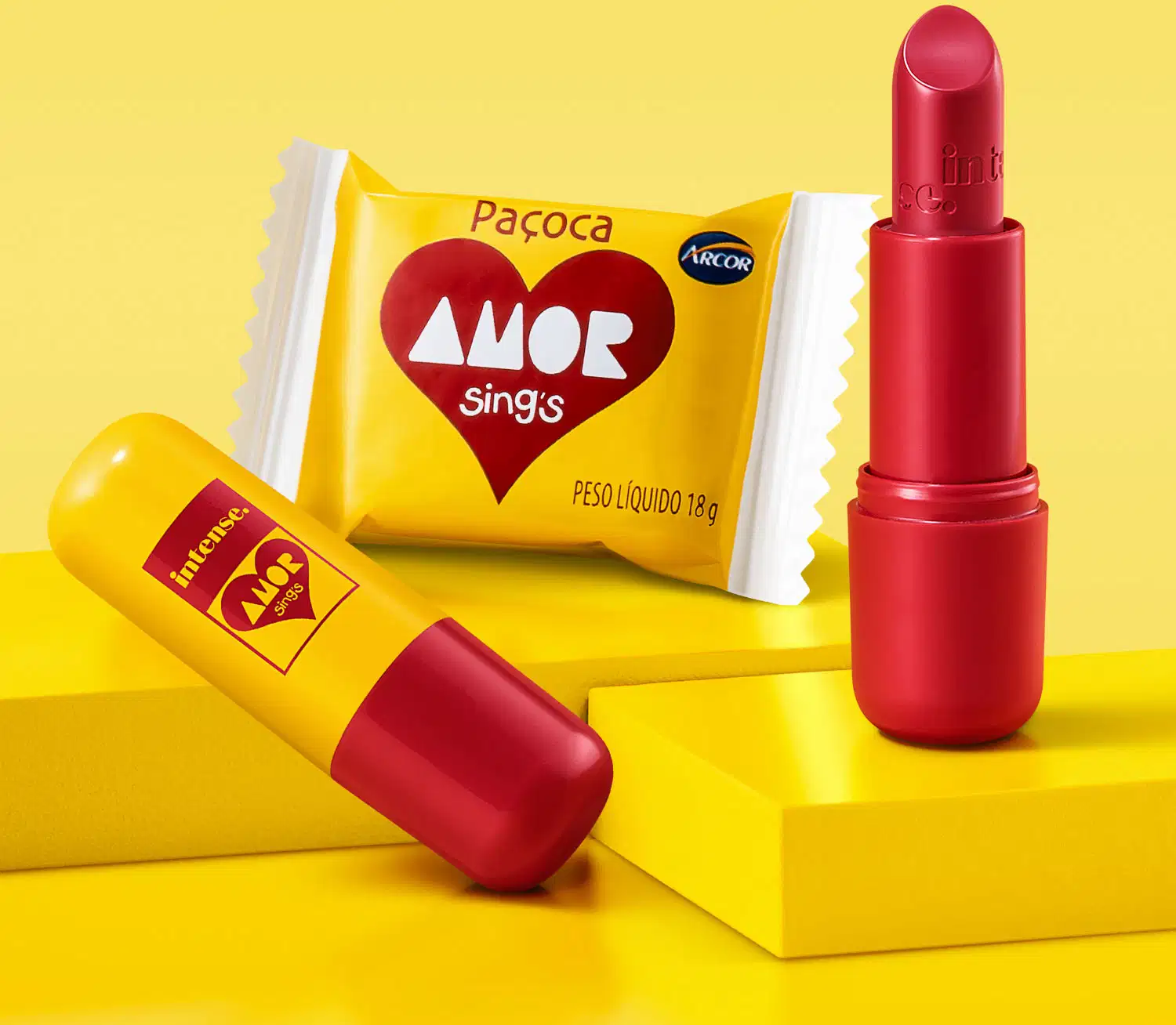 Batom cremoso vermelho da marca Intense By Amor de tampa amarela com tubo e letras vermelhas em cenário amarelo com doce ao redor.