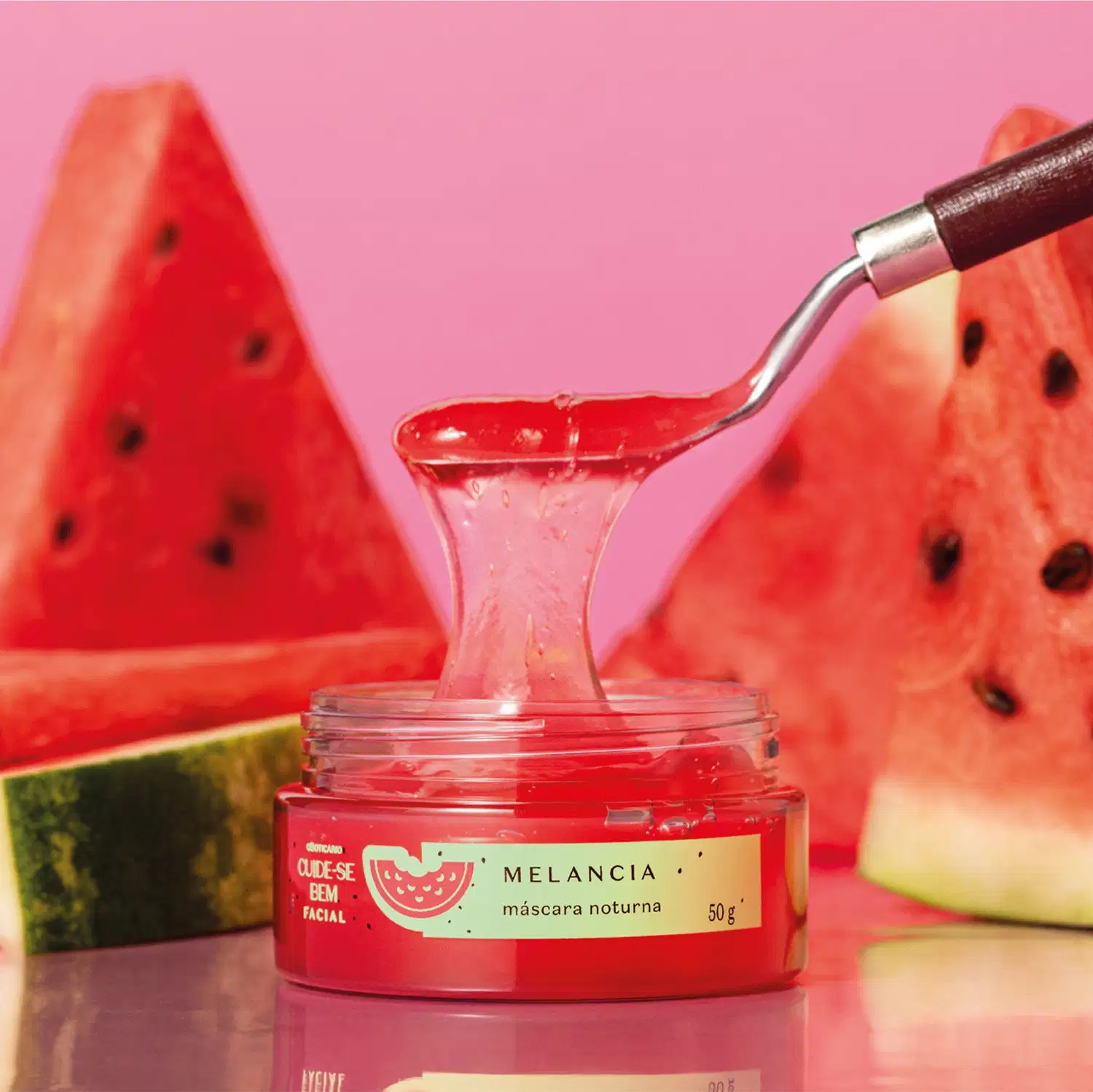 Pote com creme vermelho, tampa verde e rótulo também verde com melancia ilustrada contendo máscara facial noturna Cuide-se Bem Feira Melancia sobre superfície rosa, com fatias de melancia vermelha ao redor e uma colher com um pouco do creme suspensa.