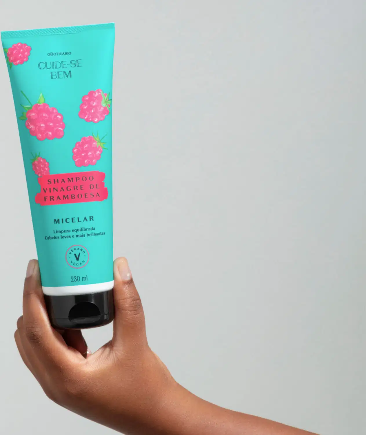 Mão segurando um frasco azul com tampa preta e ilustrações de framboesas na cor rosa contendo shampoo Cuide-se Bem Feira Vinagre de Framboesa em fundo cinza claro.