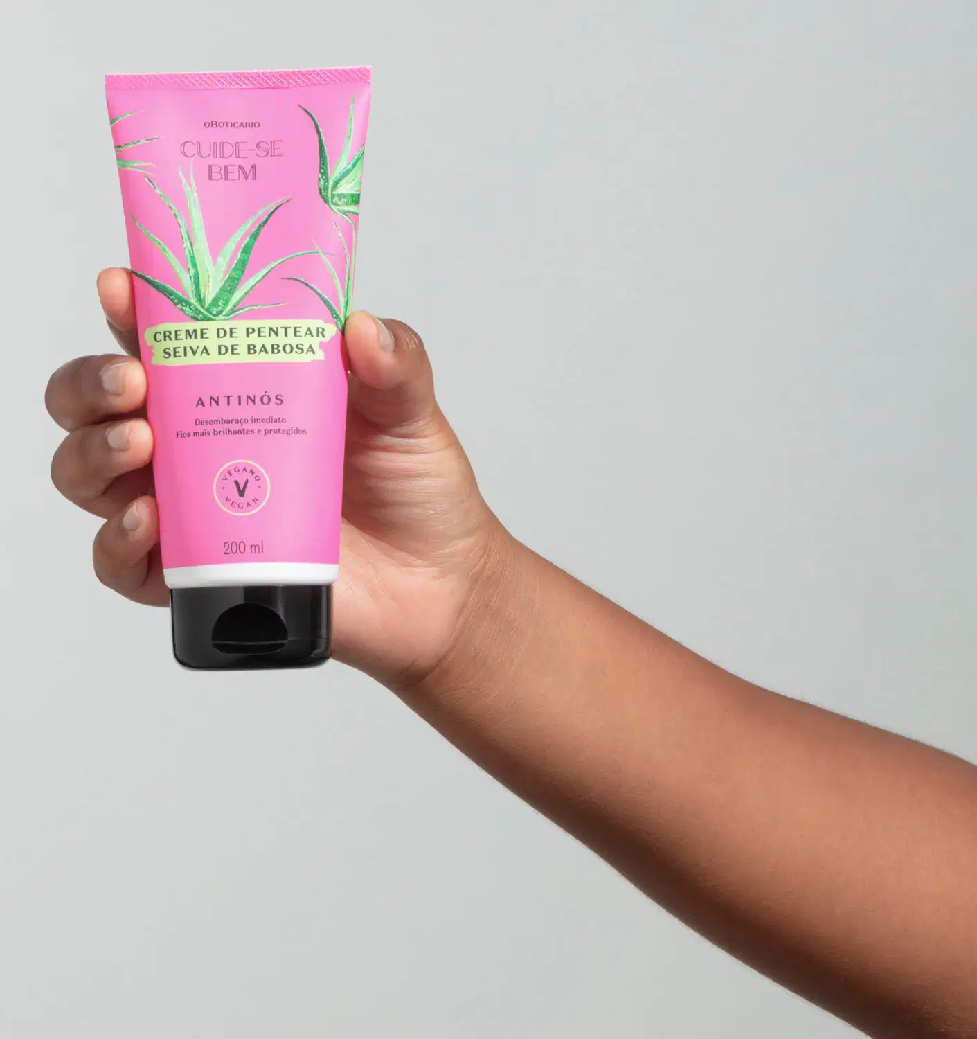 Mão segurando um frasco na cor rosa com tampa preta e ilustração de folhas de babosa contendo creme de pentear Seiva de Babosa da marca Cuide-se Bem Feira em fundo cinza.