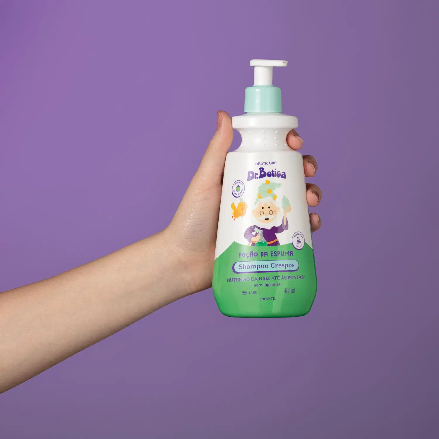 Imagem de uma mão segurando uma embalagem branca e verde com válvula dosadora azul de shampoo infantil para cabelos crespos da marca Dr. Botica em fundo azul escuro.
