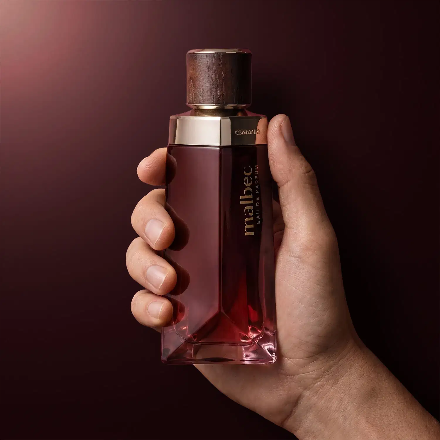 Frasco de item de perfumaria da marca Malbec em mão contra fundo vermelho escuro, com embalagem de vidro de formato geométrico e tampa de madeira.