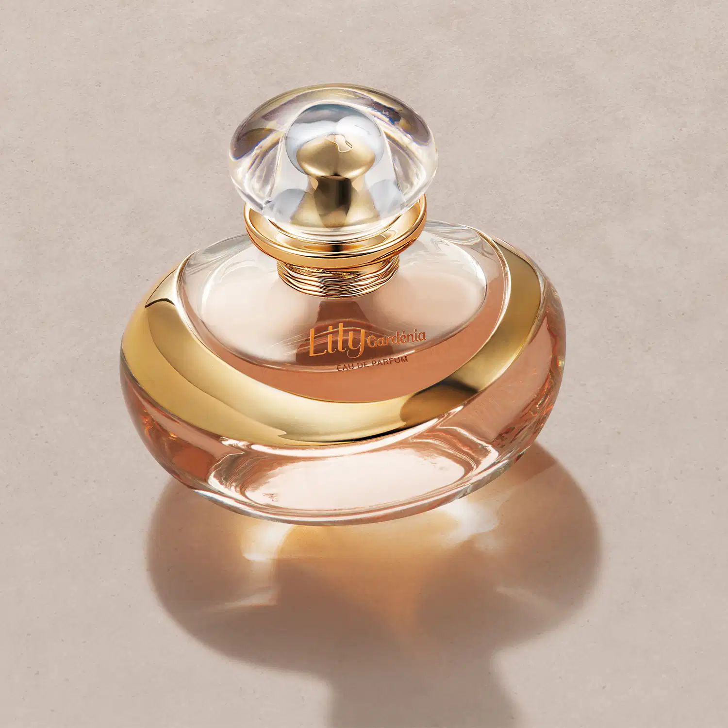O corpo do frasco apresenta um detalhe dourado com o nome "Lily Gardénia" impresso. O líquido perfumado dentro do frasco tem uma tonalidade âmbar. A base do frasco é rosa metálico e reflete a imagem, criando um efeito espelhado.