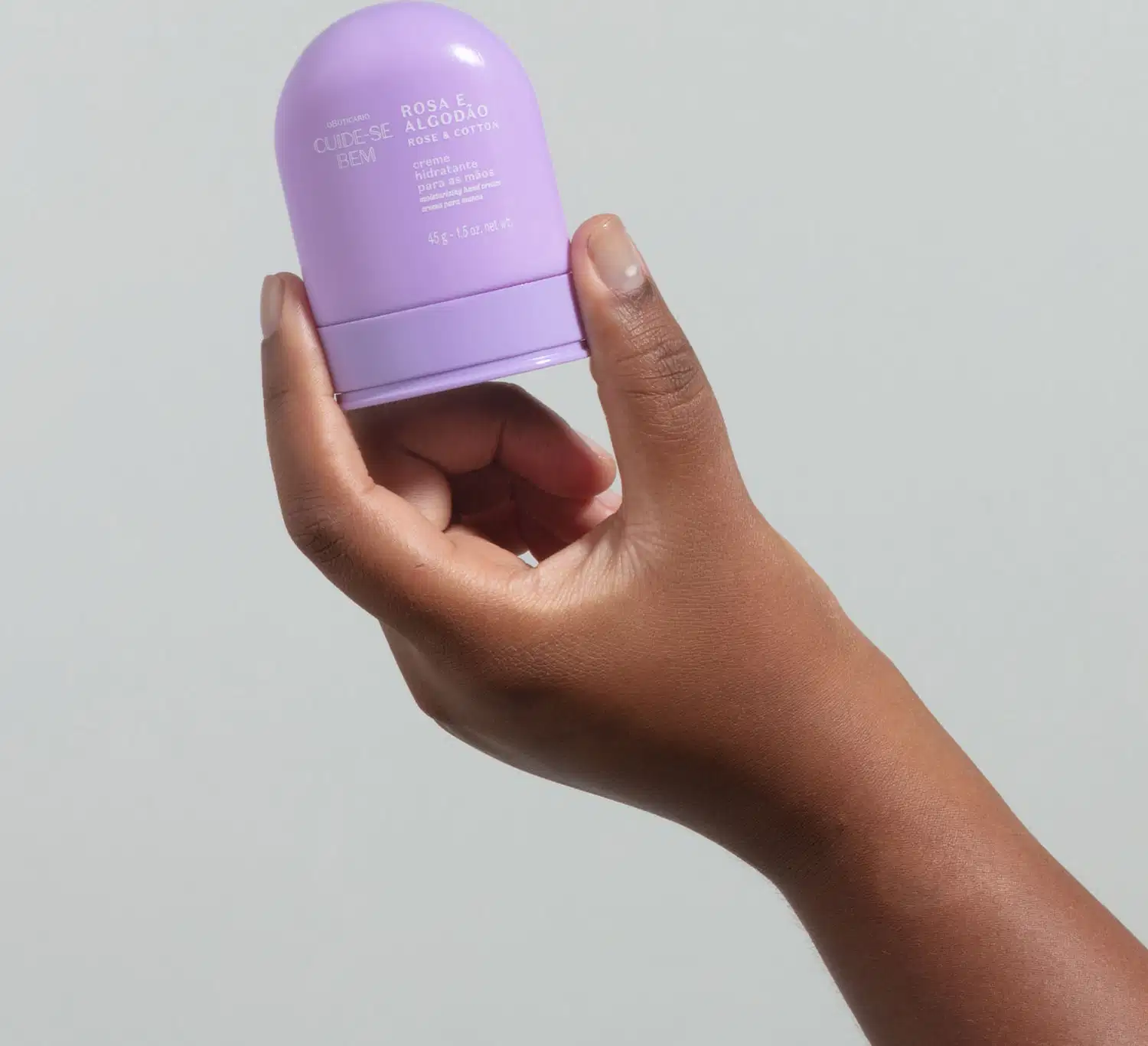 Mão segurando um Frasco roxo com cantos arredondados e letras na cor lilás contendo 45g de creme hidratante para mãos Cuide-se Bem Rosa e Algodão em fundo azul.