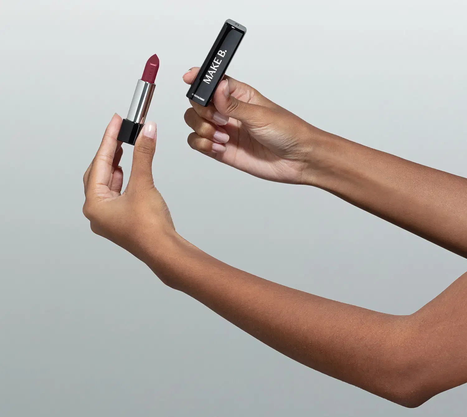 Uma mão feminina está segurando um Batom glossy na cor Vinho Blackbarry da marca Make B. em embalagem prata e preta e a outra mão segura a tampa na cor preta com letras prateadas. O fundo da imagem é cinza.