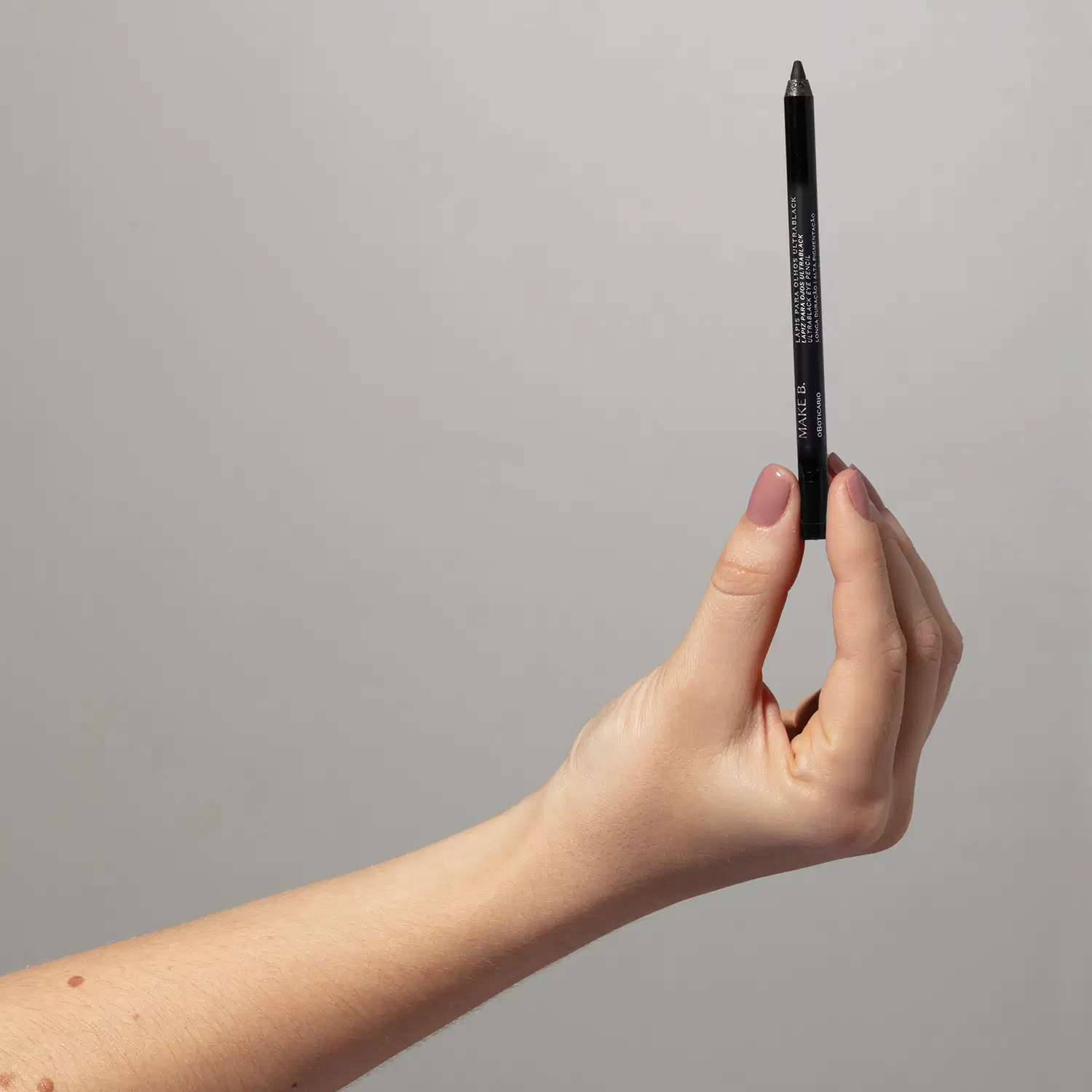 Mão feminina com unhas pintadas de lilás segurando um lápis para olhos Ultrablack Make B. em um fundo cinza.