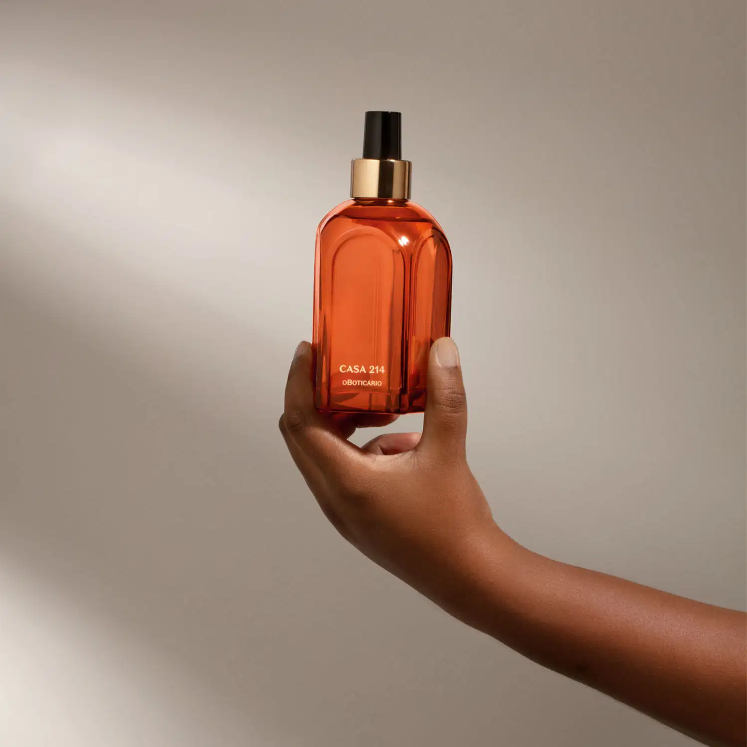 Mão segurando um vidro laranja de tampa spray preta com detalhe e letras douradas contendo aromatizador de ambientes Casa 214 Vanilla Sublime em fundo cinza com sombras.