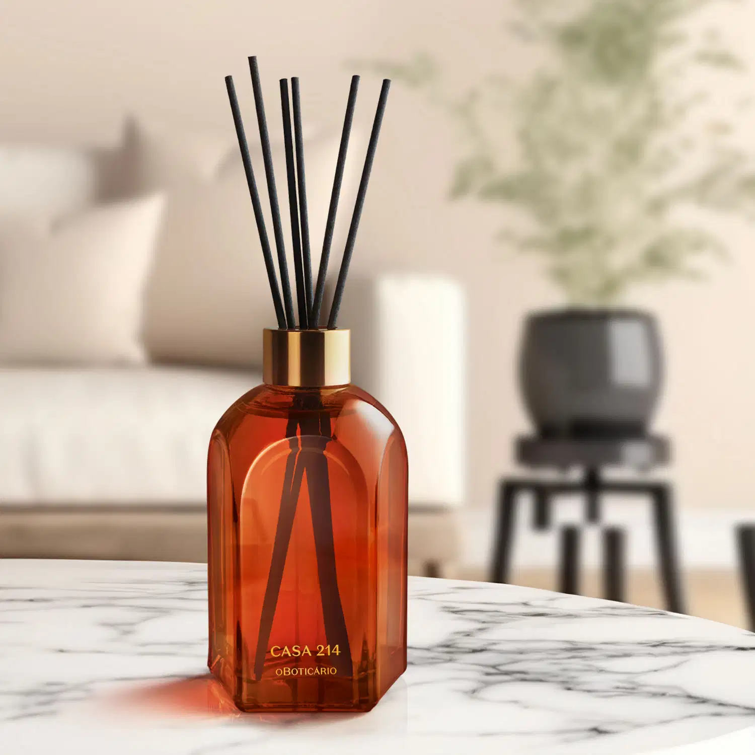 Difusor de ambientes em vidro laranja com varetas e letras douradas da marca Casa 214 Vanilla Sublime sobre superfície de mármore branco e cinza, com objeto preto desfocado ao fundo.