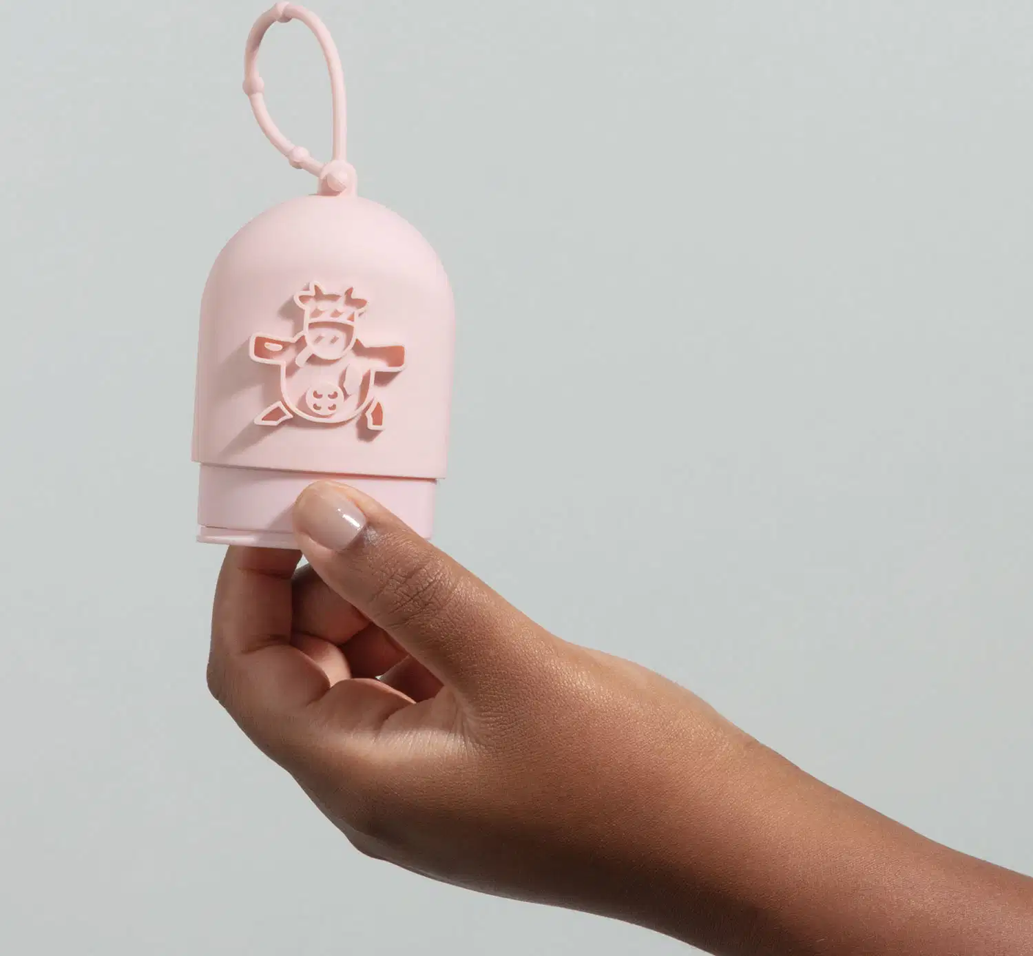 Mão segurando uma capinha de silicone rosa para creme de mãos da marca Cuide-se Bem Deleite, com ilustração de uma vaquinha em relevo e alça para pendurar. O fundo da imagem é azul claro.