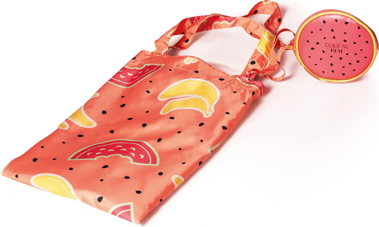 Sacola com alça de ombro e transversal na cor rosa com ilustrações de melancias, bananas e sementes pretas da marca Cuide-se Bem Feira. Acompanha um porta-moedas vermelho com pontinhos pretos, simulando melancia.