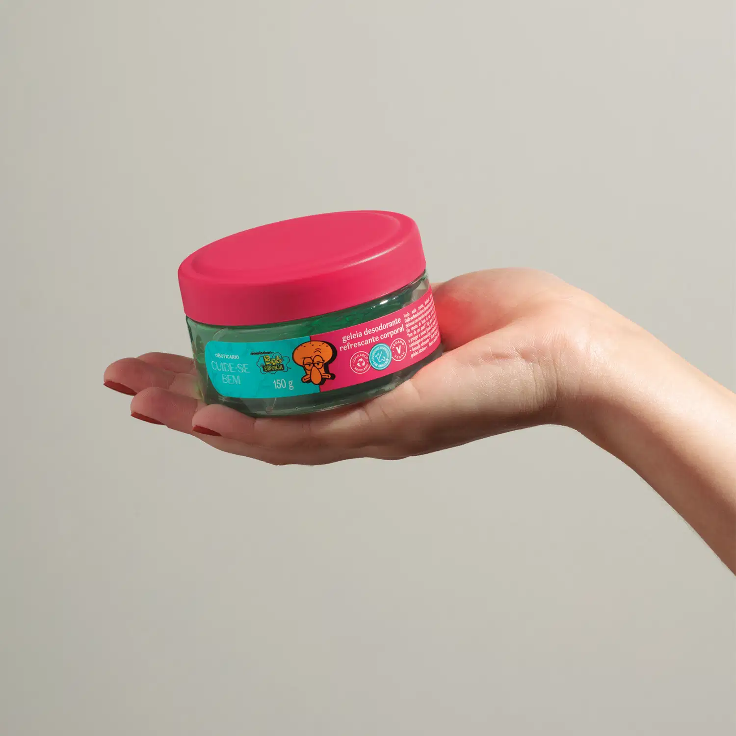 Mão segurando um pote transparente com gel verde, tampa rosa e rótulo na cor rosa e azul com ilustração do personagem Lula Molusco contendo 150g de geleia desodorante refrescante corporal da marca Cuide-se Bem Bob Esponja e Amigos em fundo cinza.