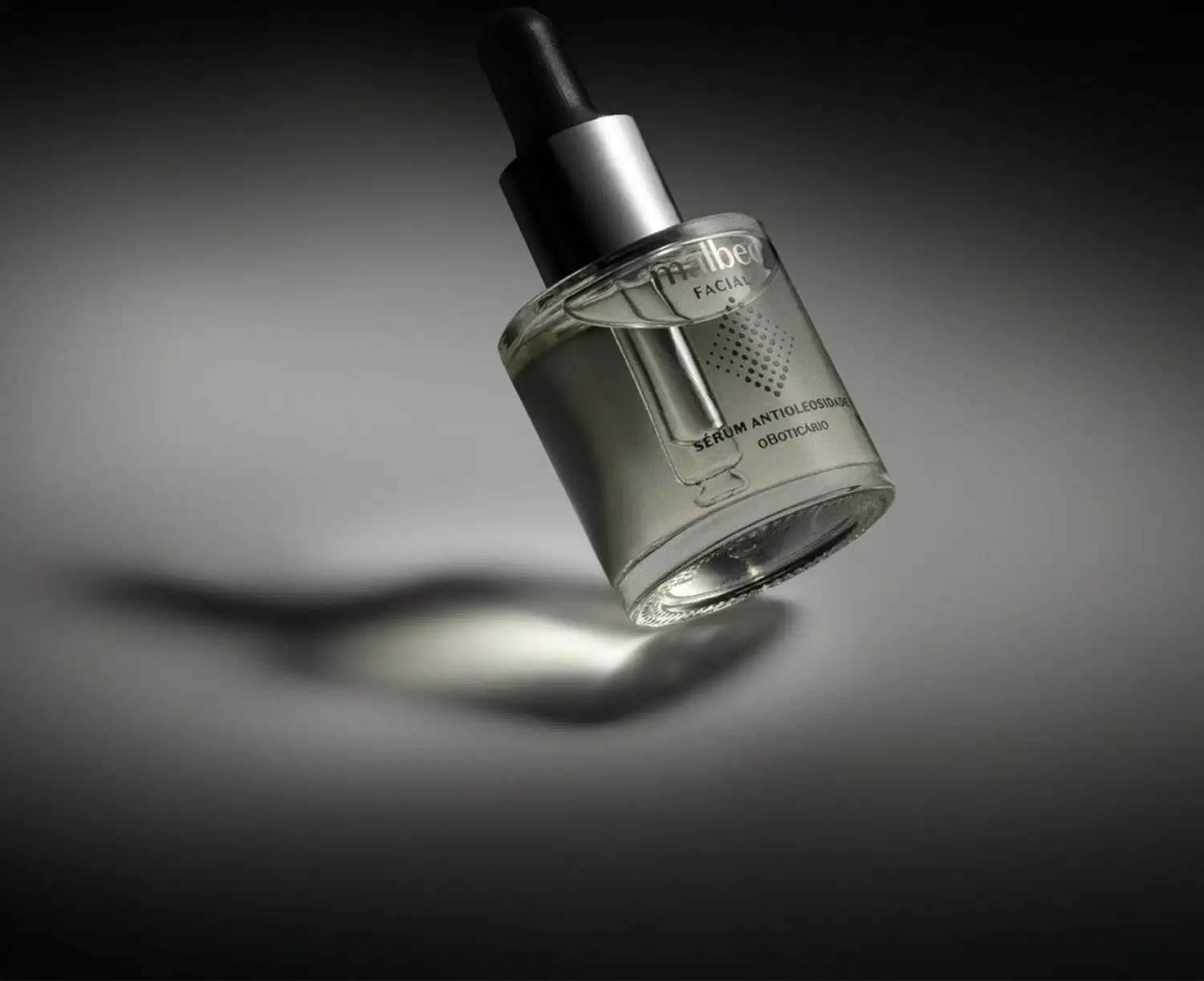 Frasco com líquido transparente, tampa prateada, conta-gotas preto e rótulo prateado de sérum antioleosidade Malbec Facial em fundo preto com iluminação em cima da embalagem.