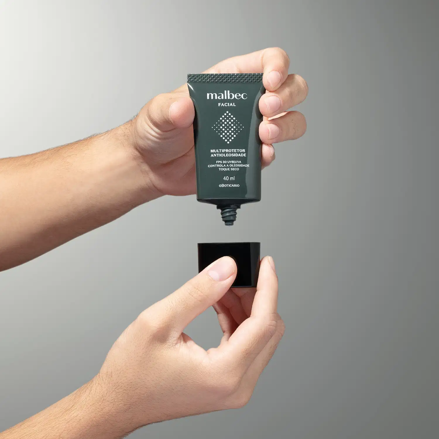 Mão masculina segurando um frasco cinza escuro com tampa preta e letras na cor cinza claro e branco de multiprotetor antioleosidade FPS30 Malbec Facial em fundo em tons de cinza.