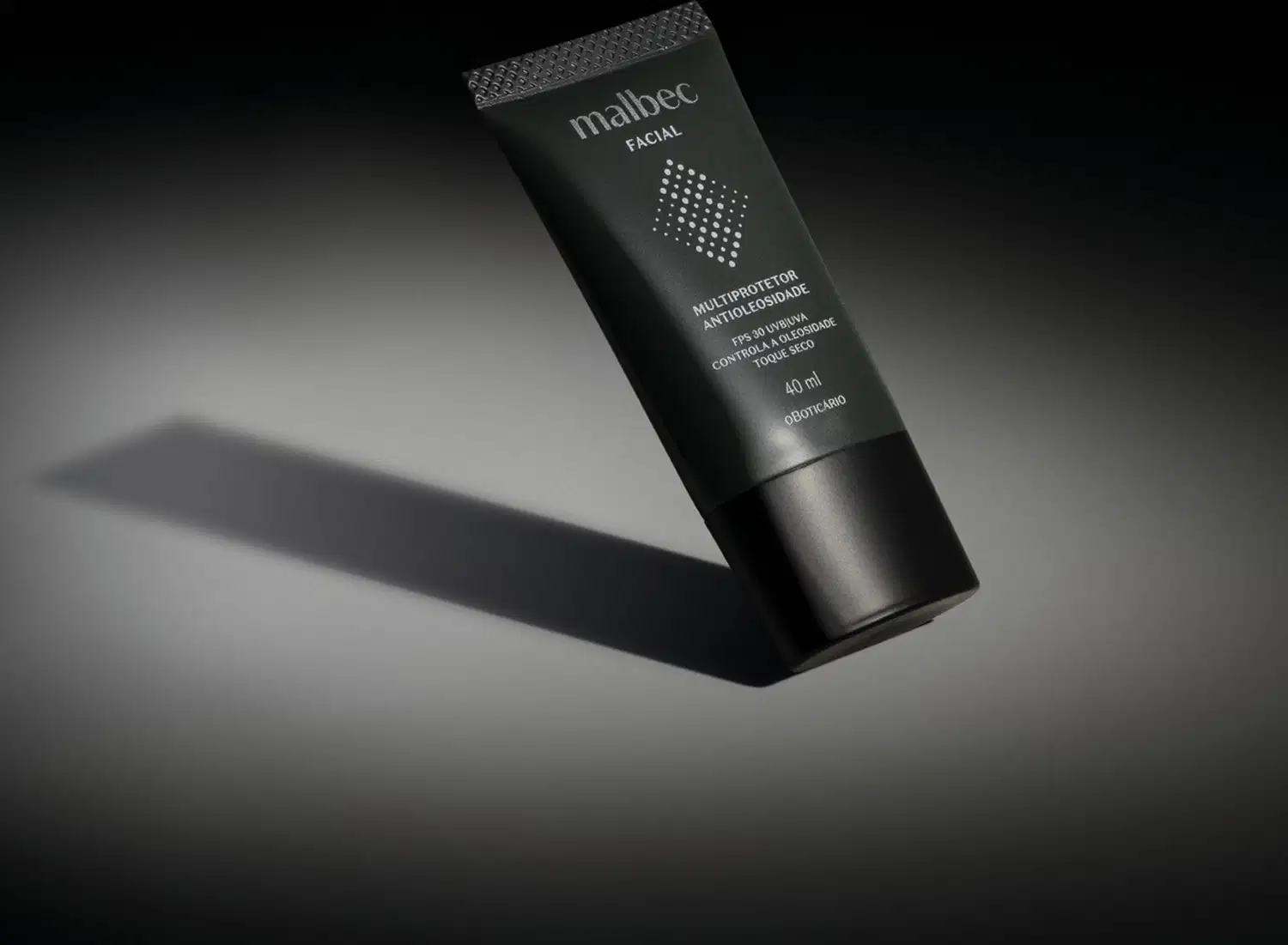 Frasco cinza escuro com tampa preta e letras na cor cinza claro e branco de multiprotetor antioleosidade FPS30 Malbec Facial em fundo preto com iluminação em cima do produto.