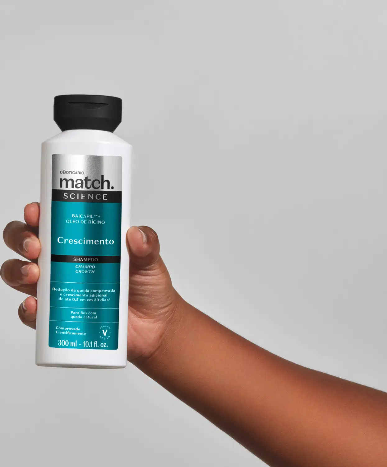 Mão segurando um vidro branco com tampa preta e rótulo azul, preto e prateado de shampoo Match Science Crescimento em fundo cinza.