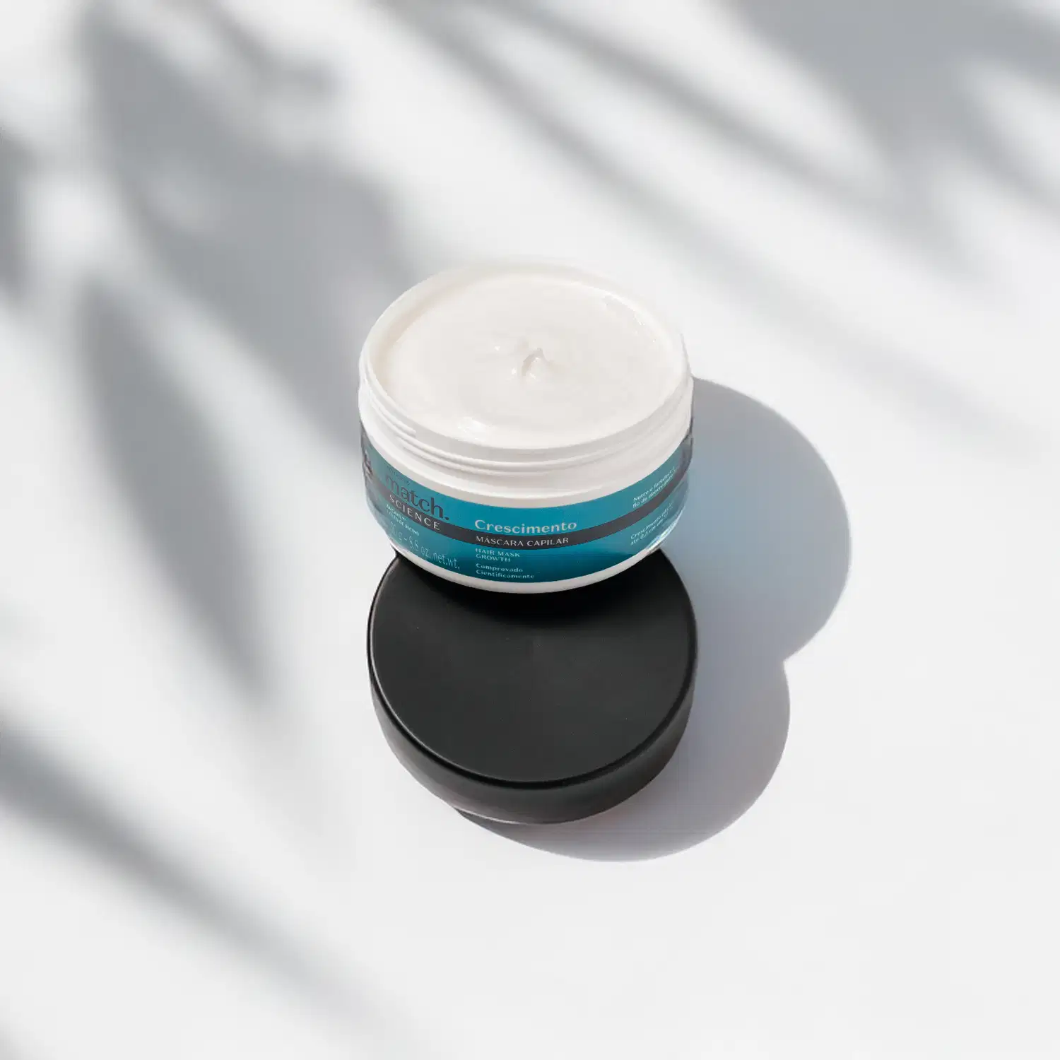 Pote branco aberto com creme branco dentro dele e tampa preta apoiada na embalagem. O produto possui rótulo na cor azul metalizado, preto e branco. A embalagem está em fundo claro com sombra de folhagens.