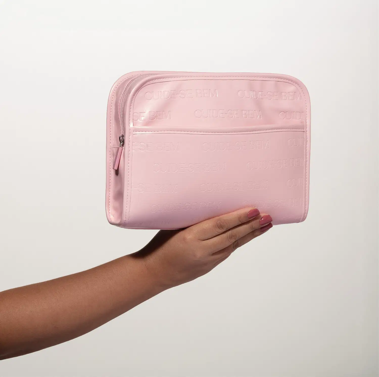 Mão feminina segurando um necessaire em material sintético na cor rosa claro. Possui formato retangular com cantos arredondados. Ele está em fundo cinza.