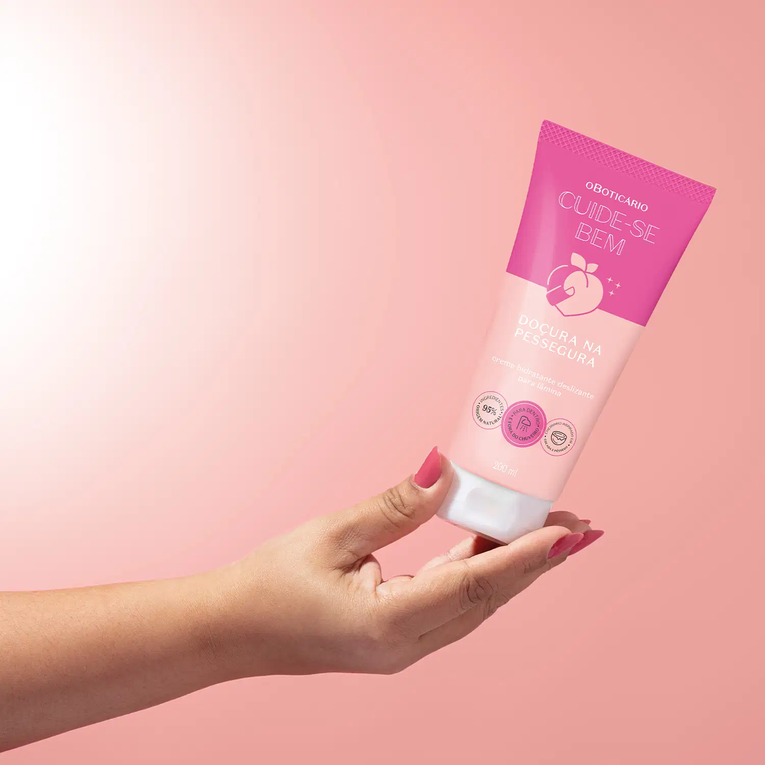 Bisnaga de creme deslizante da linha "Cuide-se Bem" da marca Boticário. A embalagem é rosa com o nome do produto "Doçura na Pessegura" impresso em letras brancas. O produto é segurado por uma mão feminina com unhas pintadas de pink contra fundo rosa.