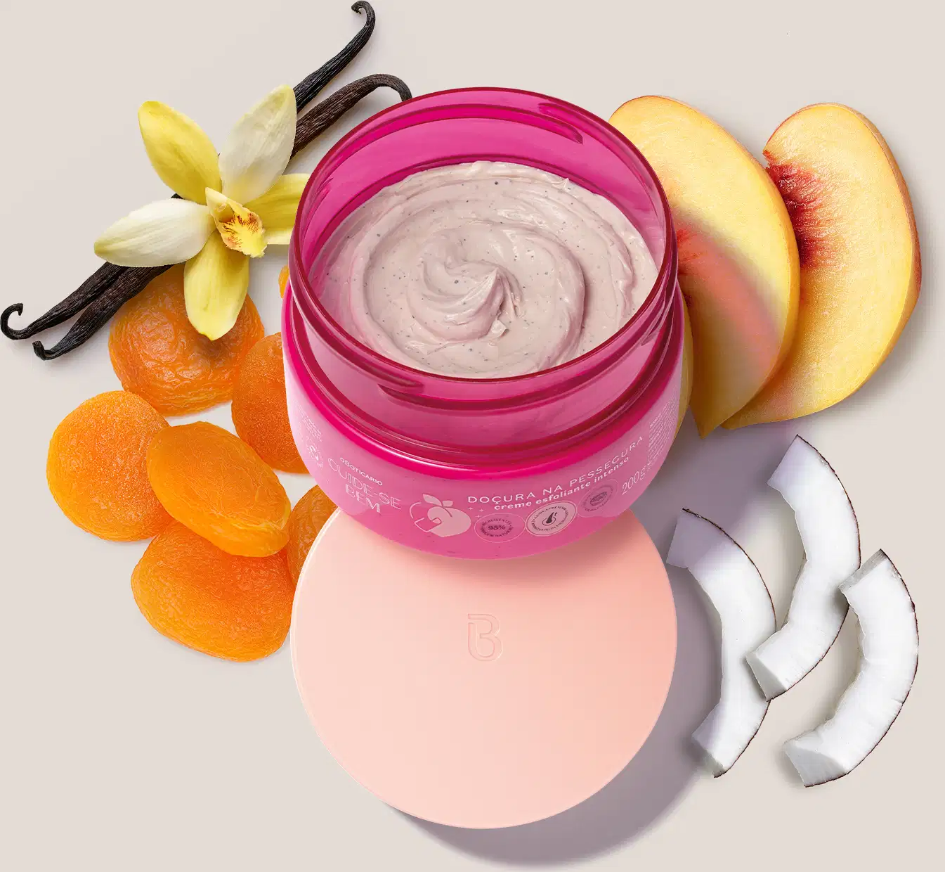 Pote pink creme esfoliante aberto com tampa rosa posicionada embaixo dele e diversos ingredientes naturais, como flores de baunilha, fatias de pêssego e rodelas de coco ao redor, sugerindo que o produto possui uma fórmula com ingredientes naturais.
